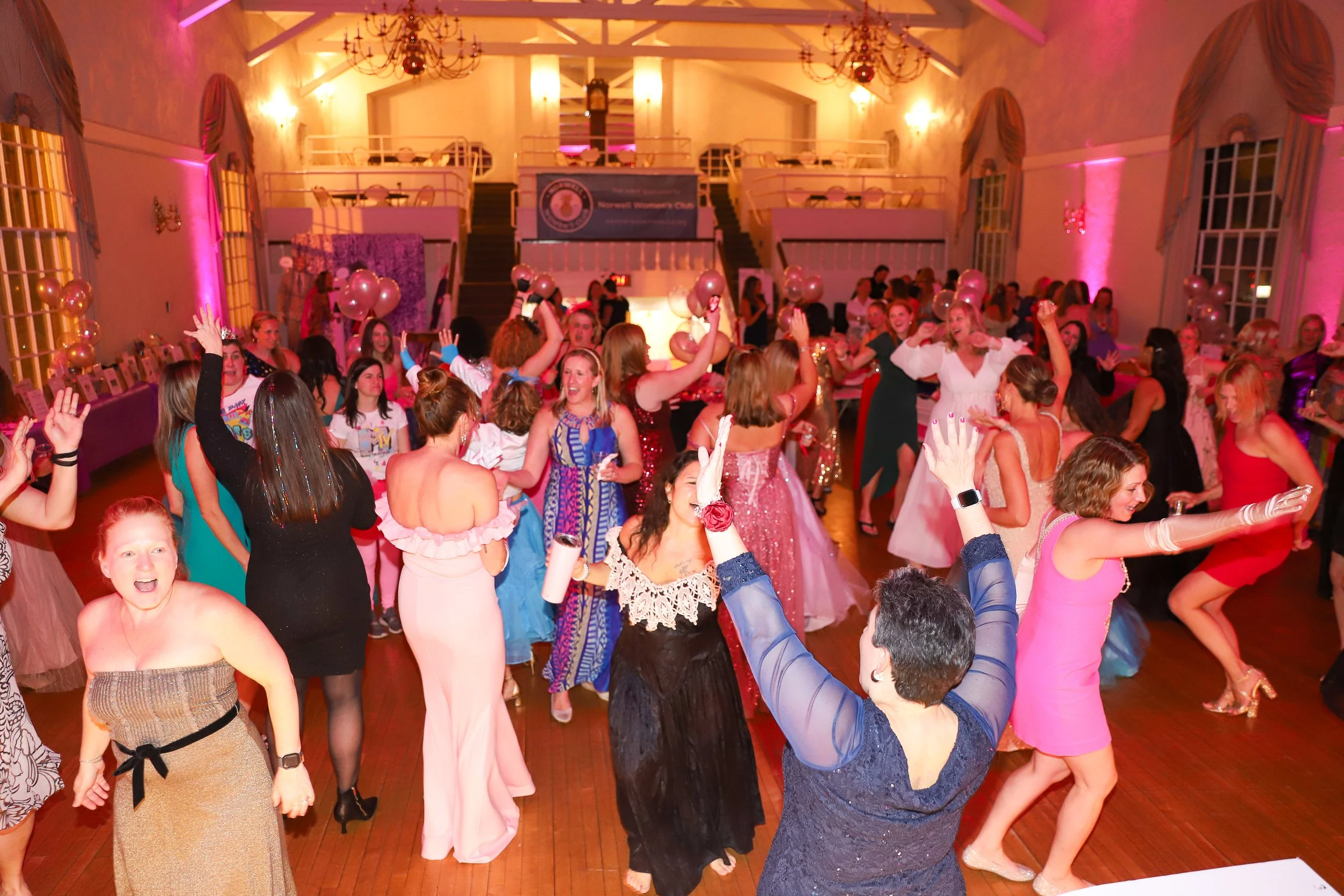 People dancing and celebrating at an indoor event, with balloons and pink lighting.