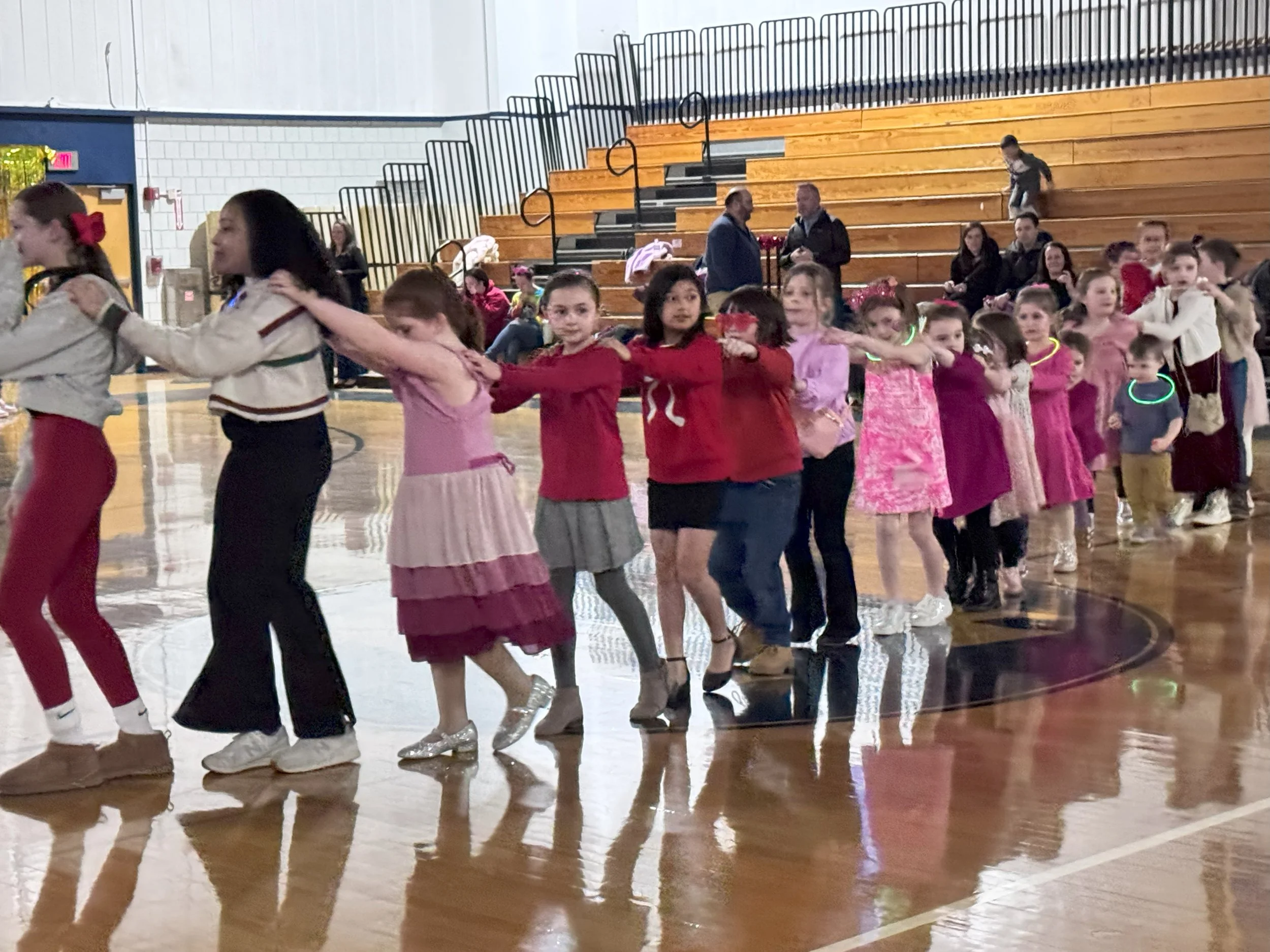 Kids in colorful clothing forming a conga line in a gymnasium with wooden bleachers in the background.