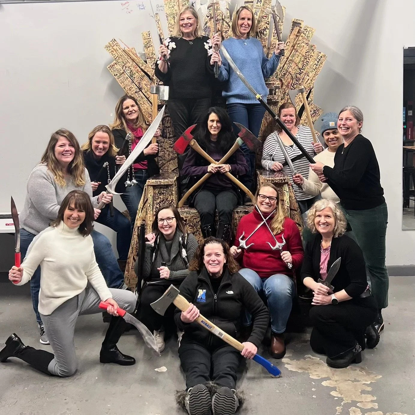 Group of women posing together with toy weapons, sitting on and around a large cardboard throne decorated with writings and symbols, in an indoor setting.