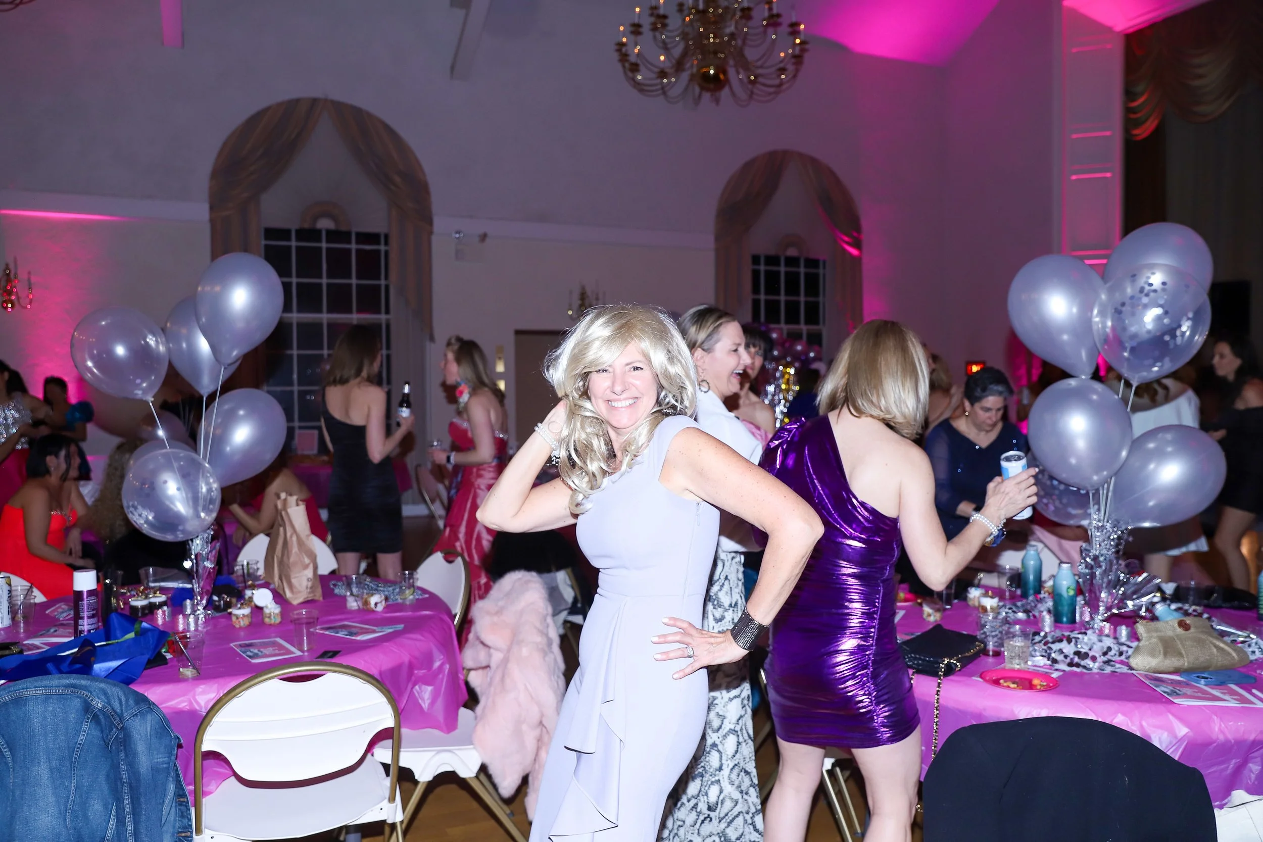 Women celebrating at a party with pink tablecloths, balloons, and women dancing and talking.
