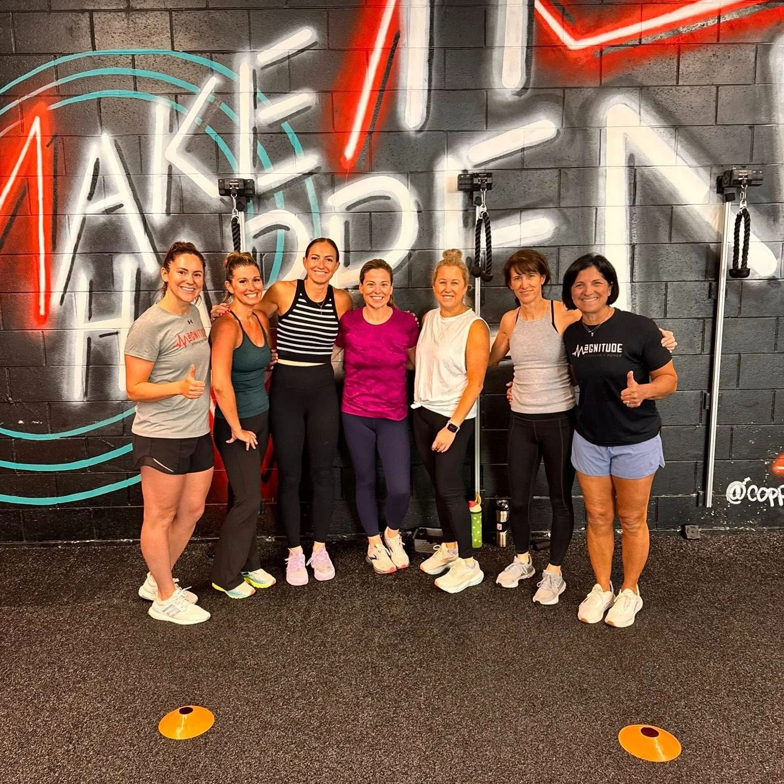 Group of seven women standing together in a fitness gym, smiling, with a neon sign on the wall behind them.