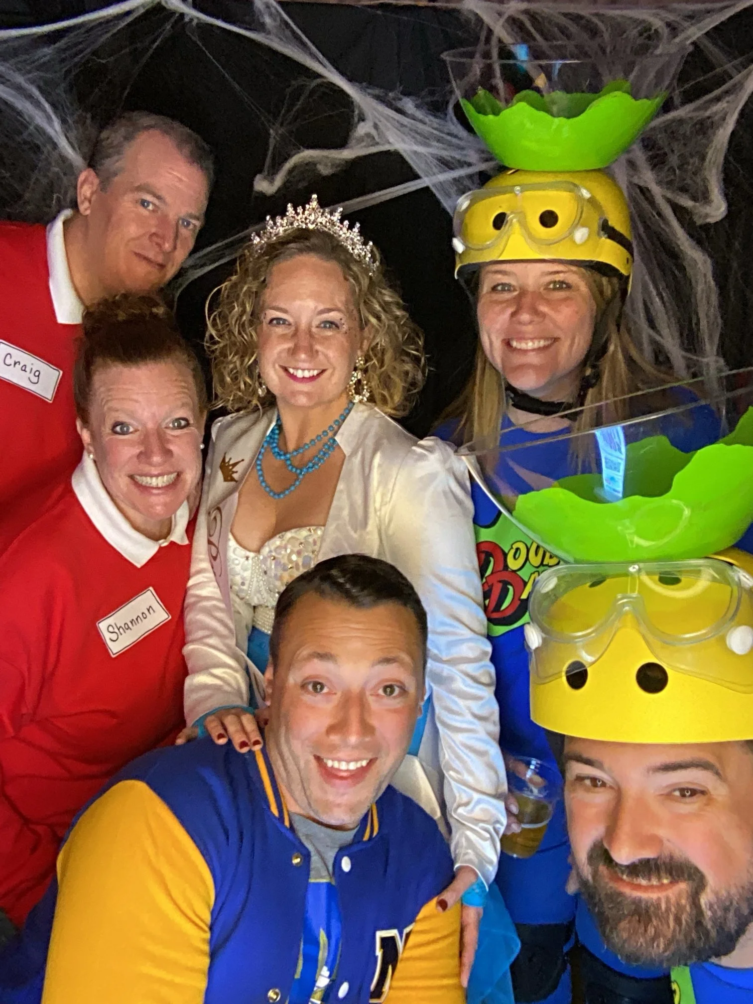 Group of six people dressed in costumes, with some wearing helmets, glasses, and crowns, smiling for a group selfie in a decorated setting with spider webs.