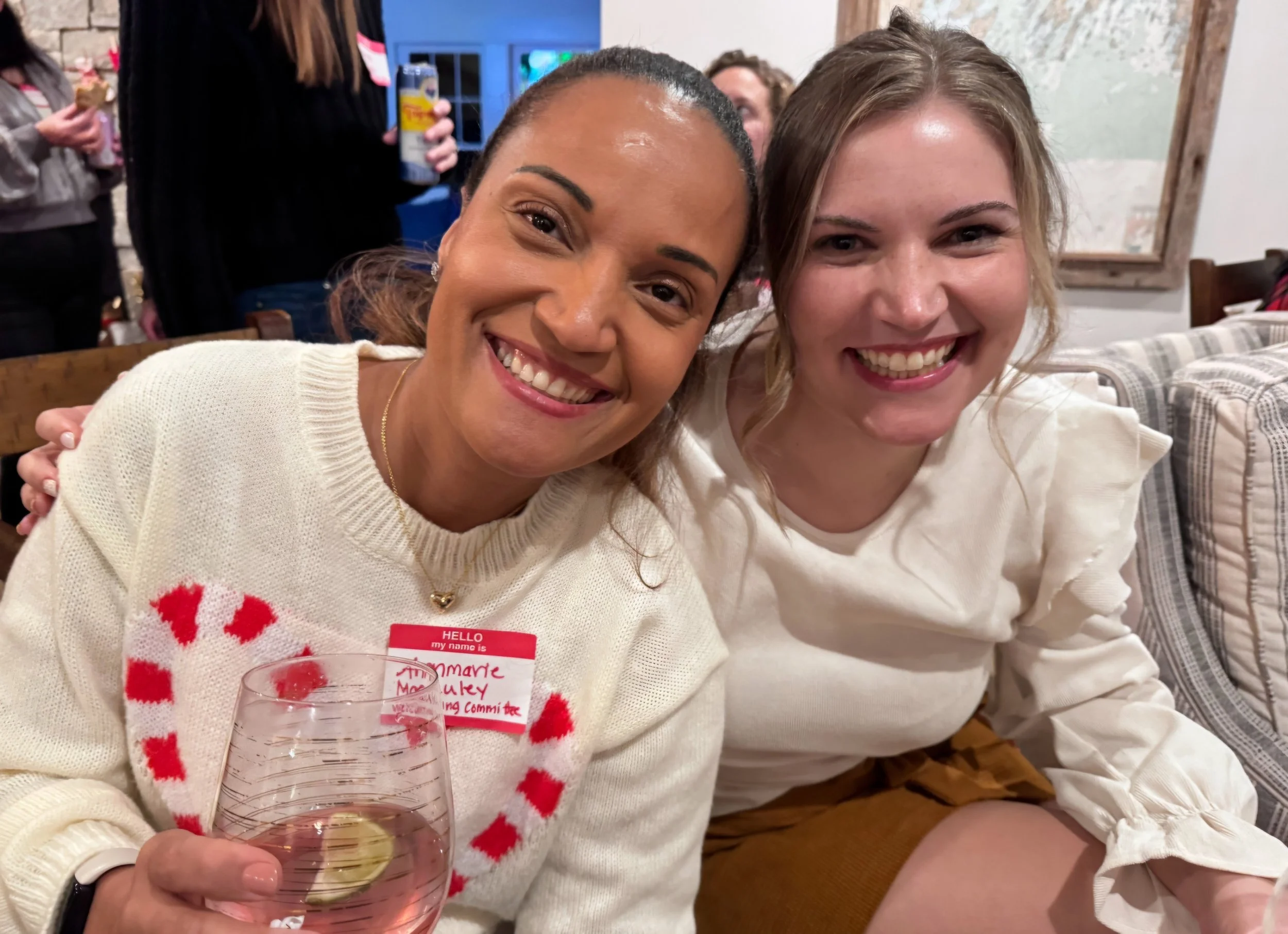 Two smiling women sitting close together at a social gathering, one holding a glass of wine with a lemon slice, both wearing white tops and enjoying each other's company.