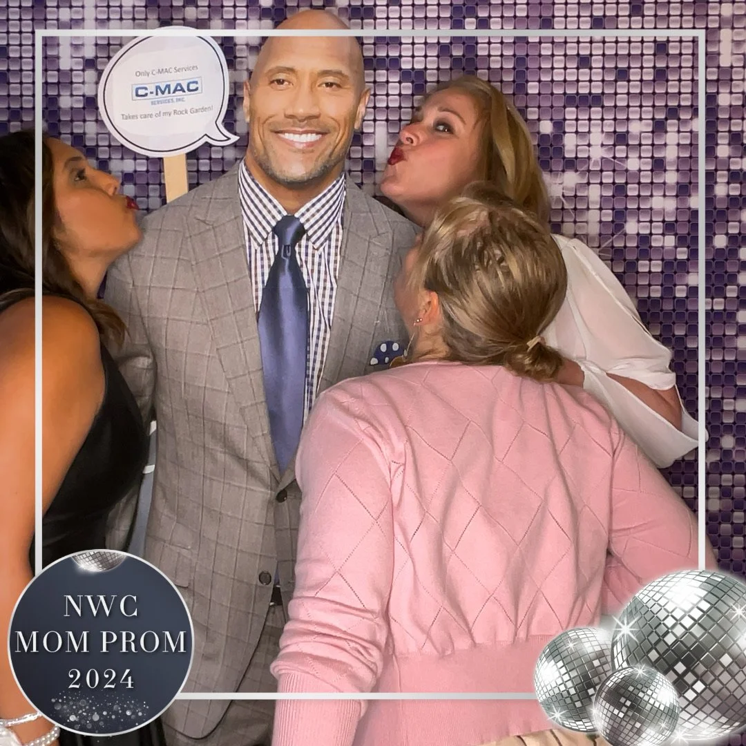 A group of four women and a man at a prom event, with one woman kissing the man on the cheek, another woman pouting near his face, and two women close to him. The man is smiling, wearing a plaid suit and tie. The background is purple with a pattern,