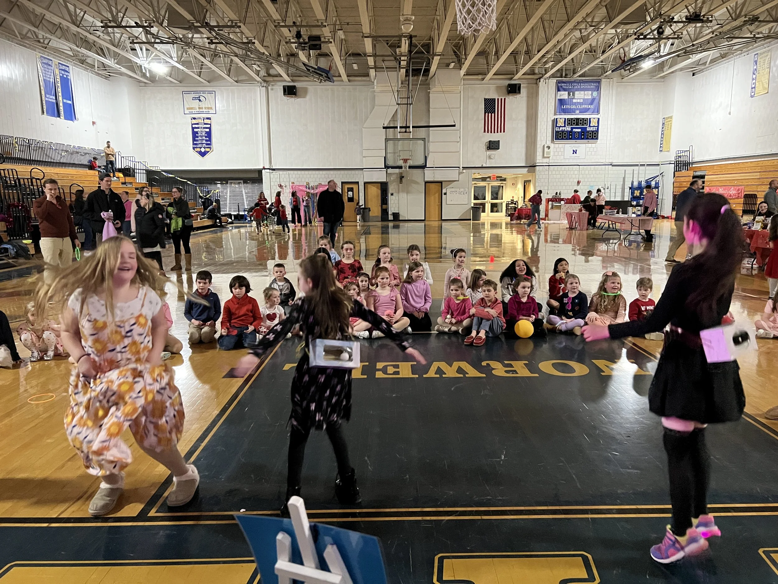 Children and adults gathered in a gymnasium for a school event or assembly, with three young girls performing in the foreground and the audience seated on the floor behind them.