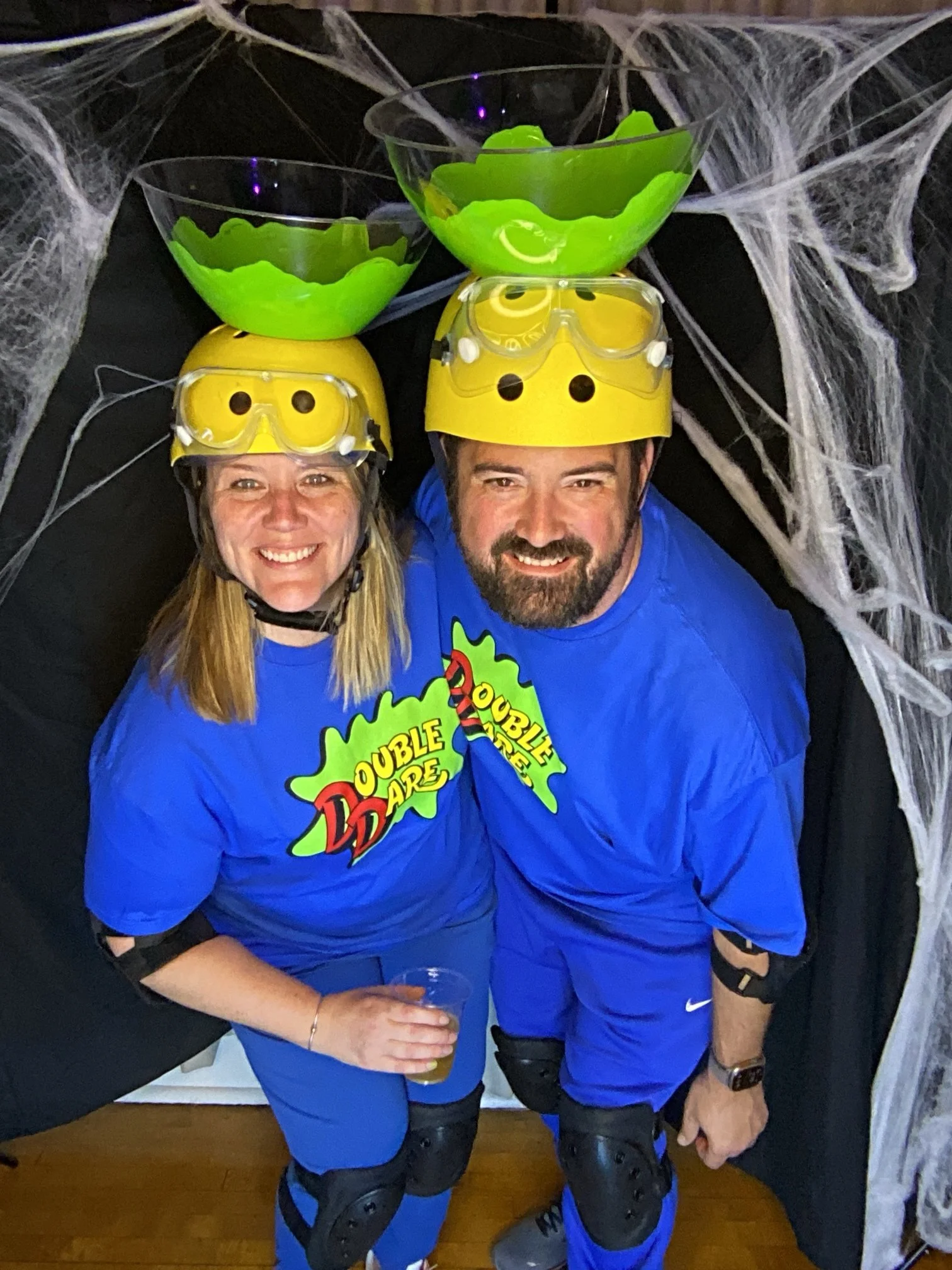 Two people dressed in blue shirts with 'Double Dare' logos, wearing yellow helmets and goggles, standing under a Halloween spider web decoration. They have bowls on their helmets filled with green and yellow slime, and are smiling at the camera.
