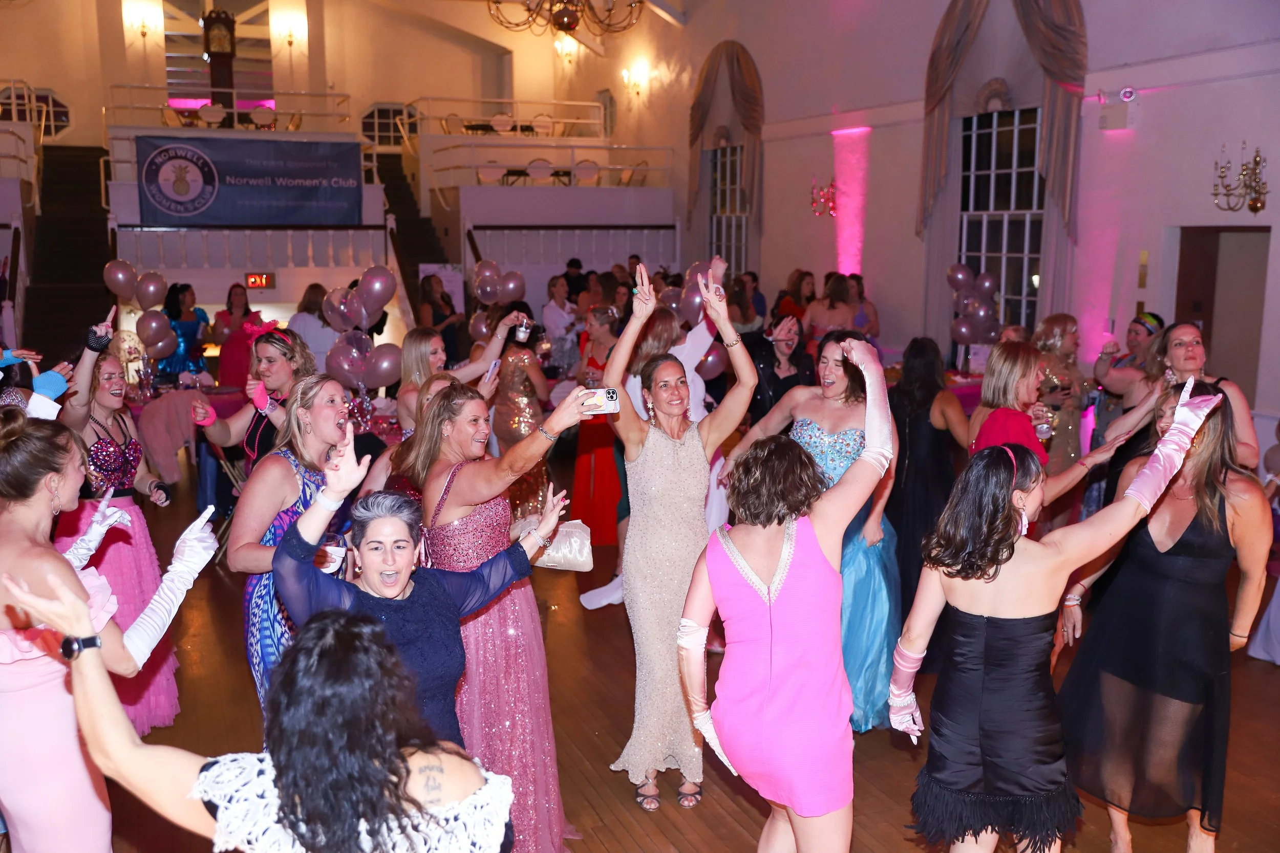 Women dancing and celebrating at a party or event with pink balloons and elegant dresses.