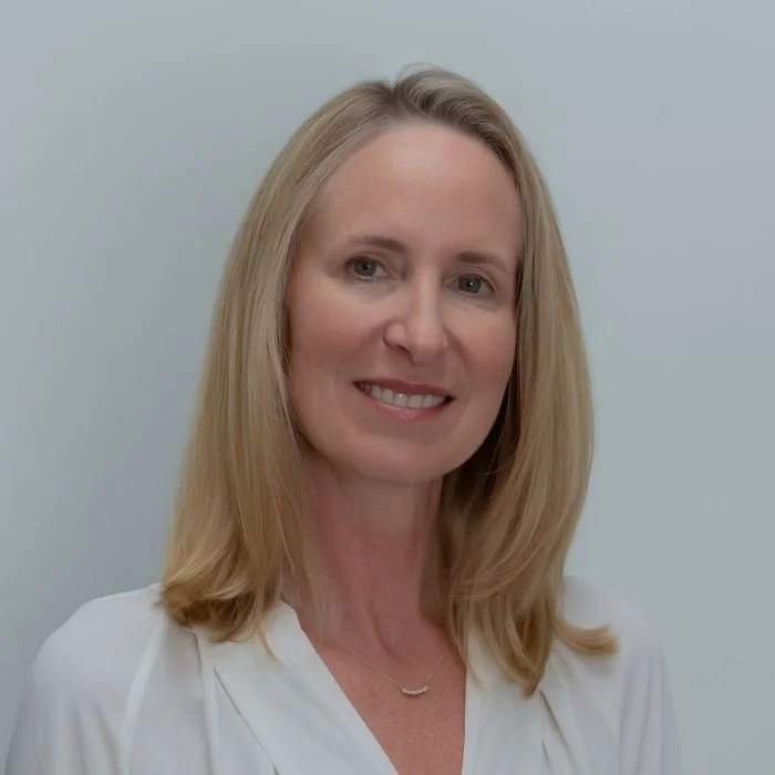 A middle-aged woman with long blonde hair, wearing a light blue top, pearl necklace, and earrings, smiling outdoors.