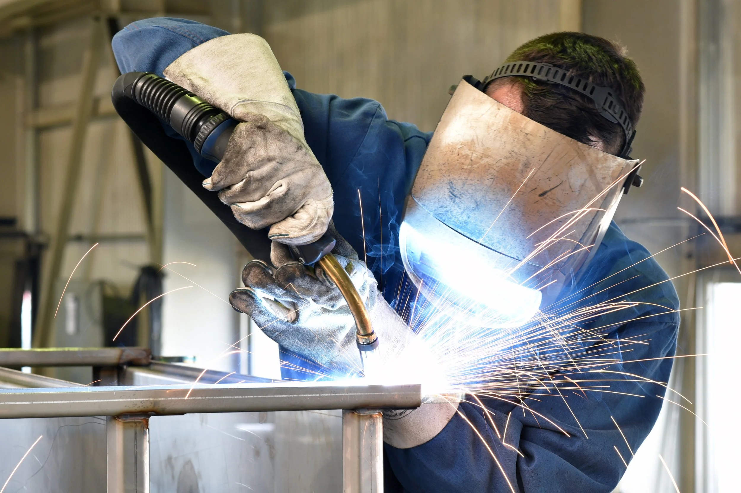 Welder wearing a protective helmet and gloves, working on metal with sparks flying.
