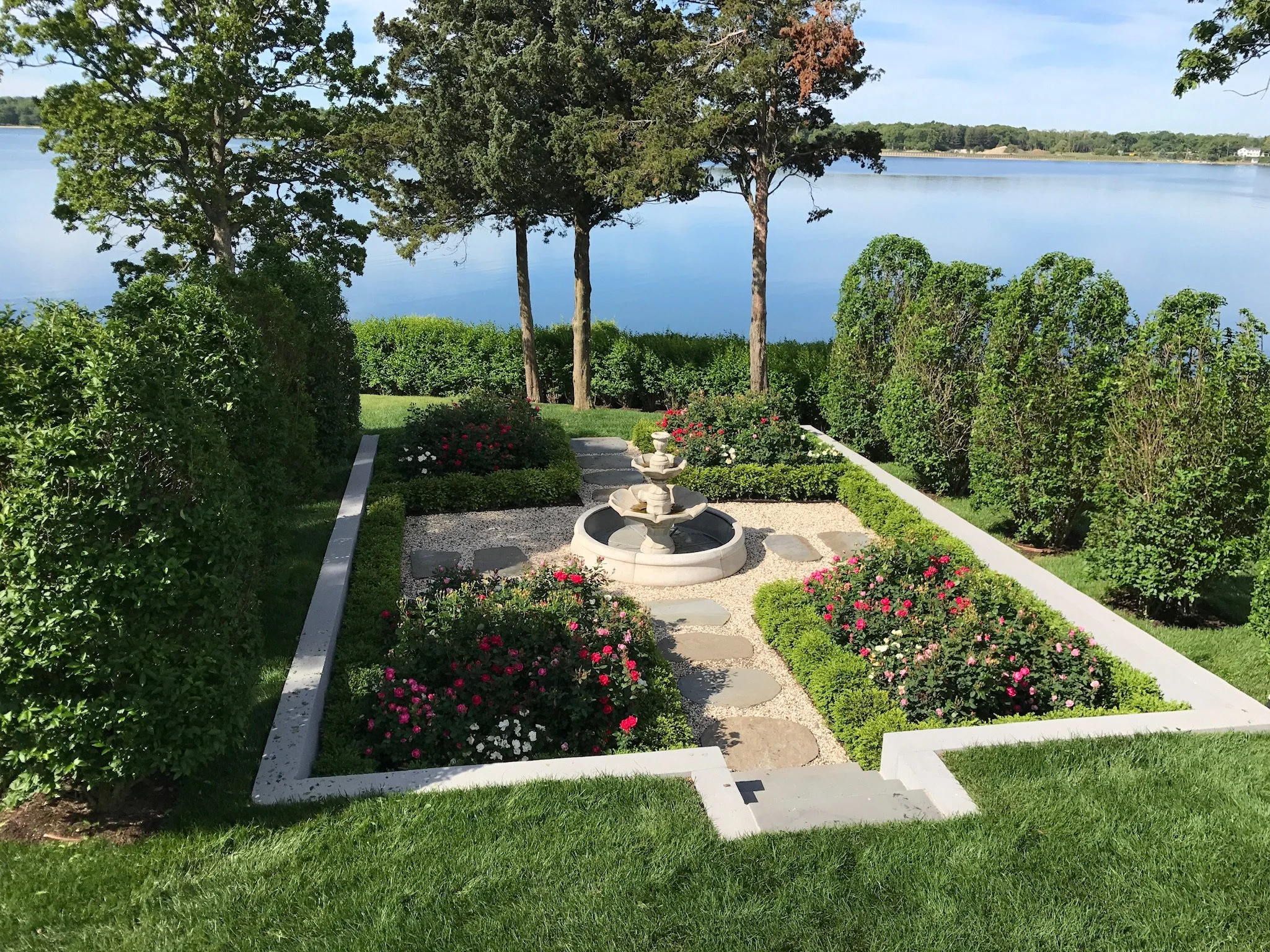 An image of a pristine rose garden with a fountain in the center overlooking a body of water.