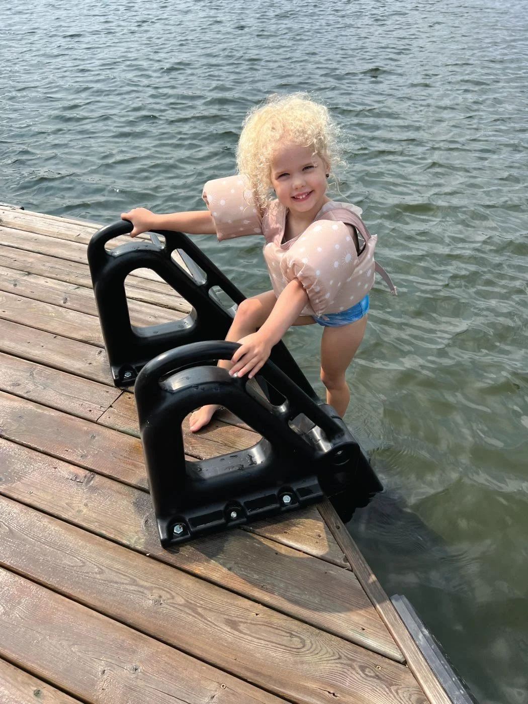 A young girl with curly blonde hair standing on a wooden dock by the water, holding a black boat cleat, smiling at the camera.