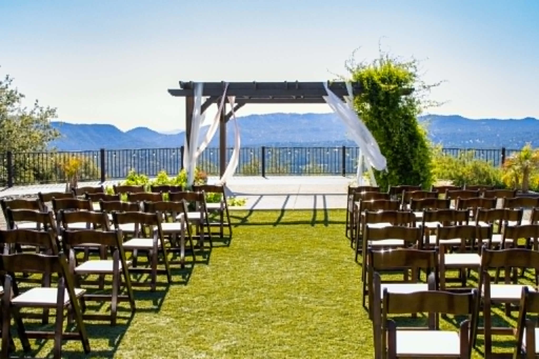 Outdoor wedding ceremony setup with rows of chairs facing an arbor with white fabric drapes, mountain view background, green grass, and trees.
