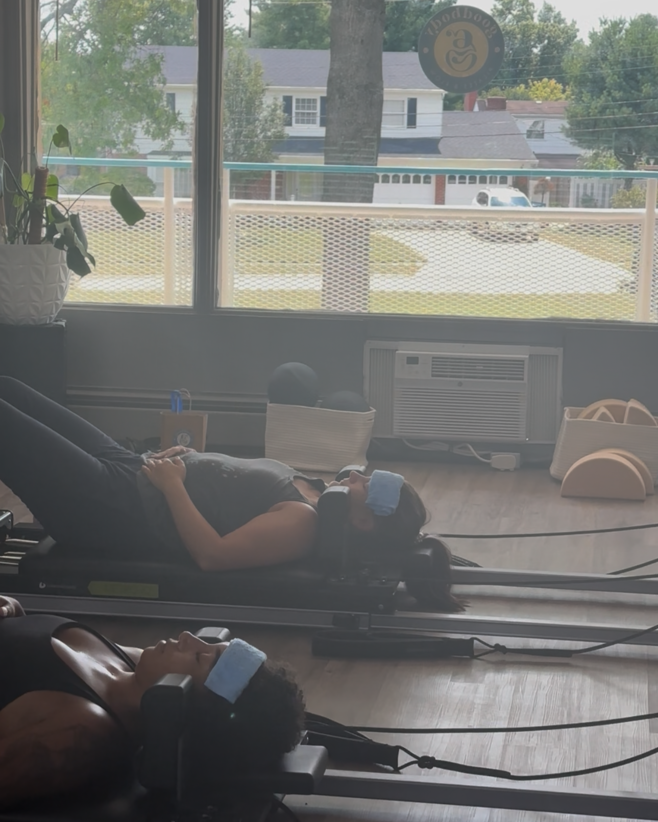 Two women performing leg exercises on reformer Pilates machines in a studio with large front window and a view of neighborhood houses and trees outside.