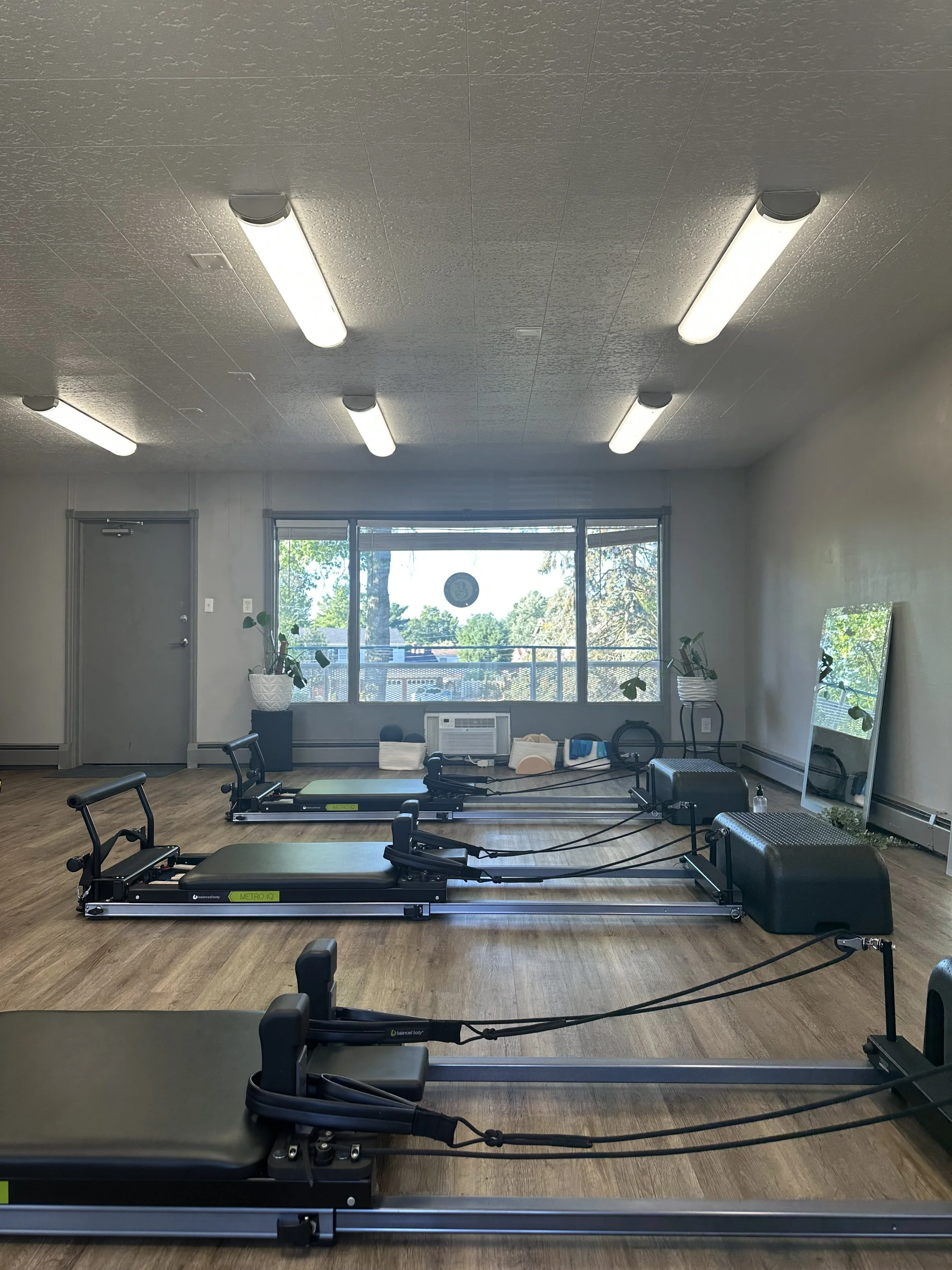 Four Pilates reformer machines lined up in a fitness studio, with a mirror and large window showing trees outside.