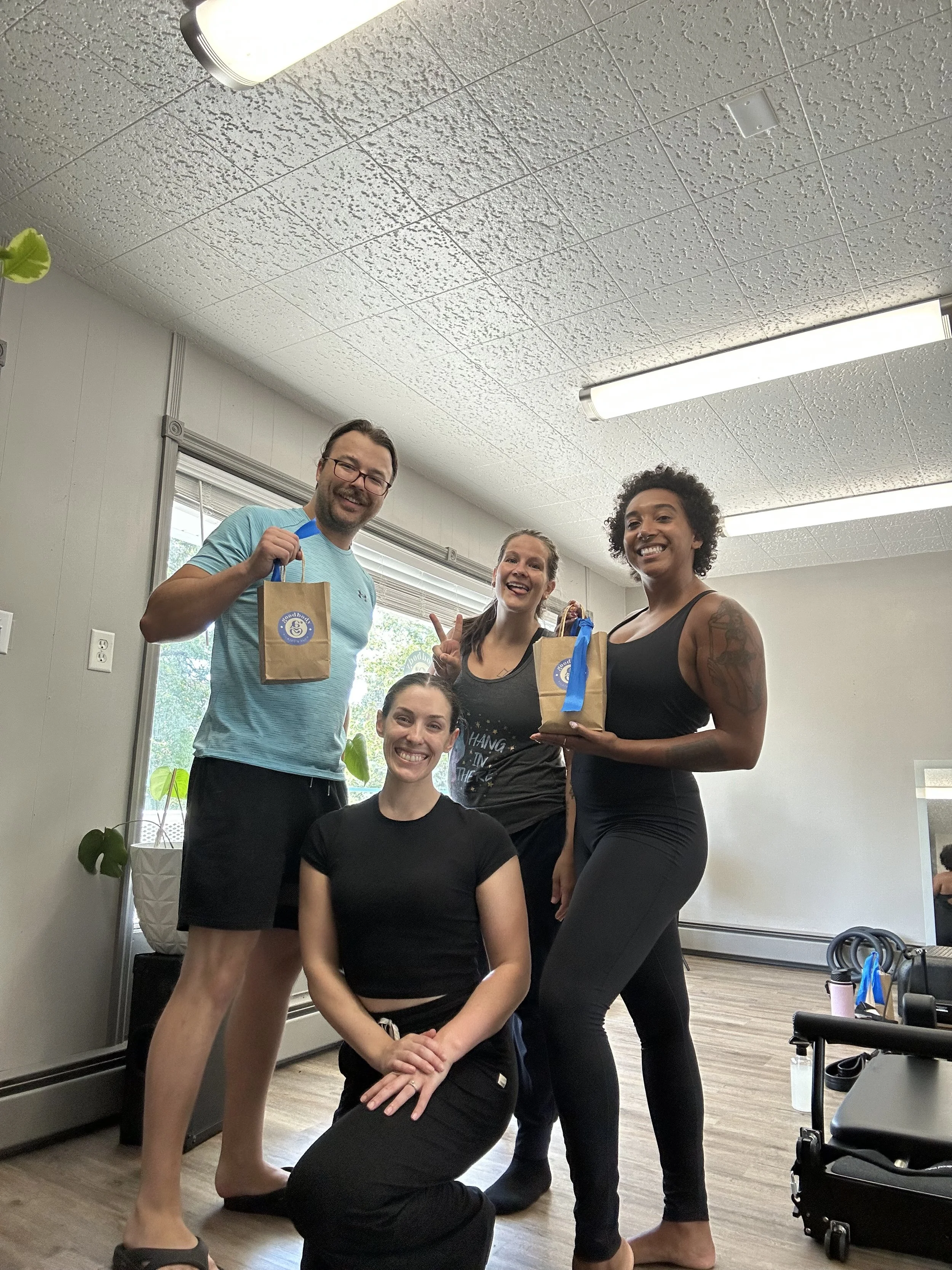 Four women and one man smiling indoors, one woman kneeling in front, two women holding gift bags, in a room with a window, plants, and exercise equipment.