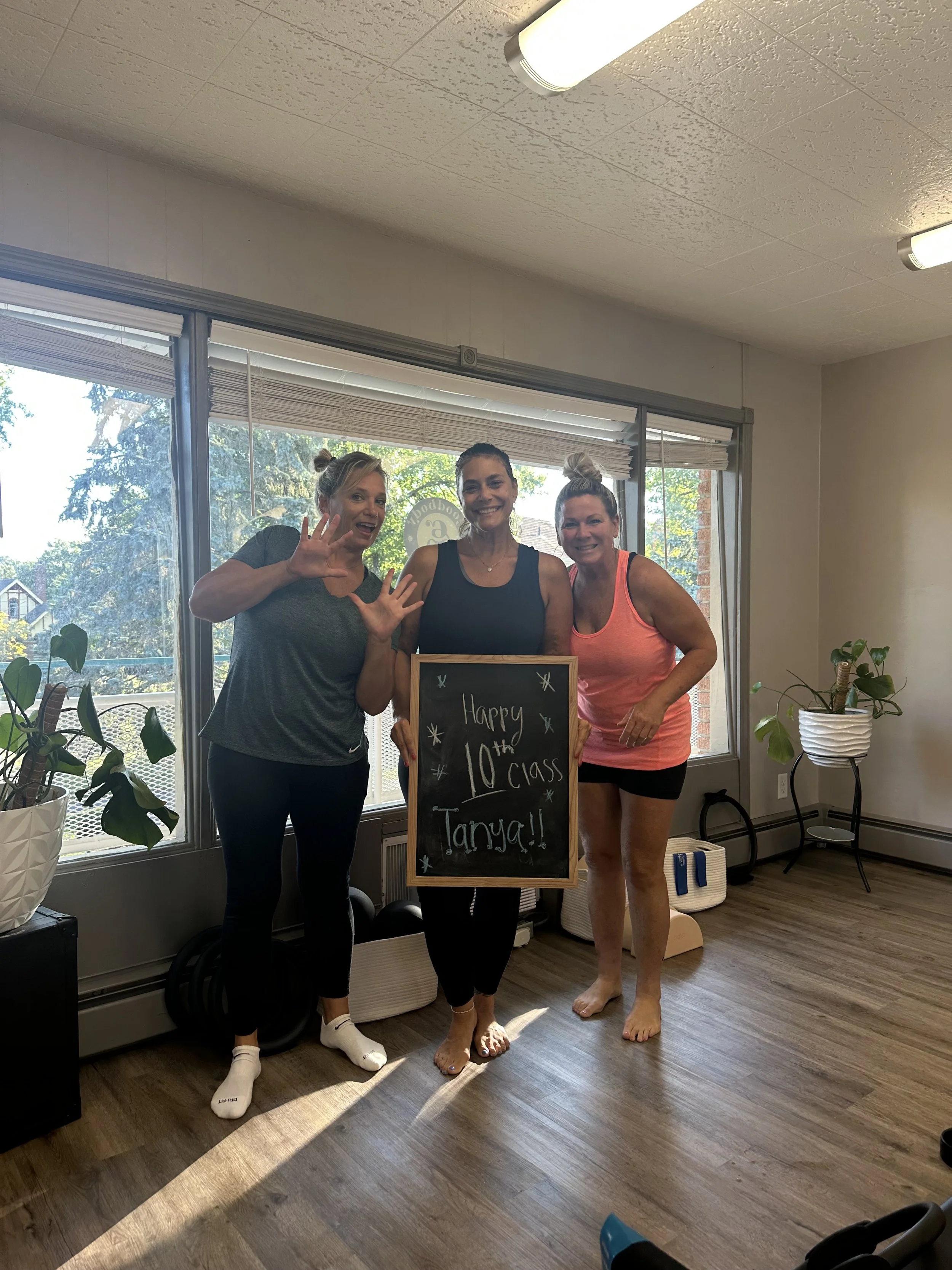 Three women celebrating a 10th class graduation, holding a chalkboard sign that says 'Happy 10th Class Tanya!!' in a brightly lit room with large windows, plants, and hardwood floors.