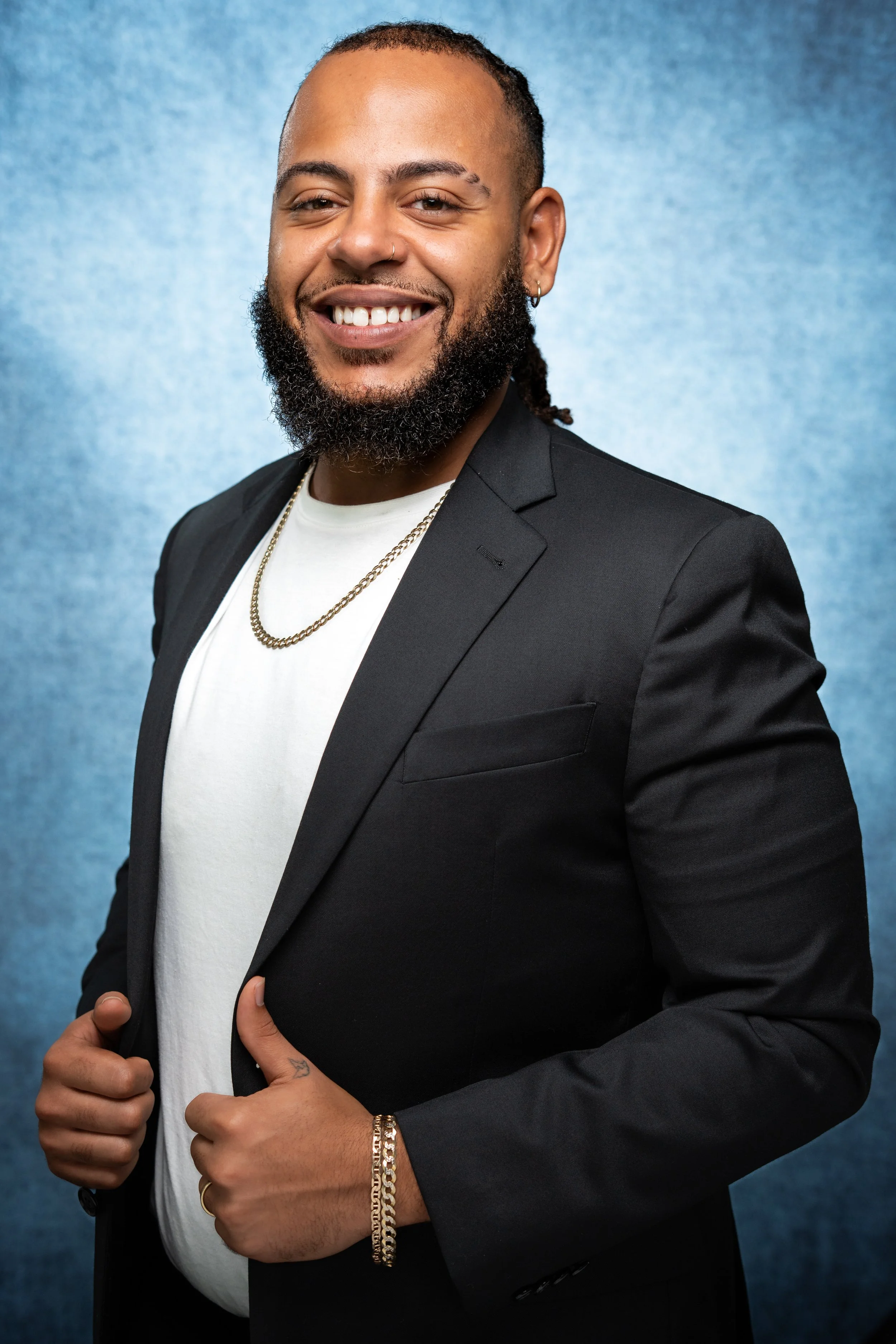 Professional picture, blue background, multi-racial man, nice beard, big smile, piercing, hand tattoo, dreads, black suit, white shirt