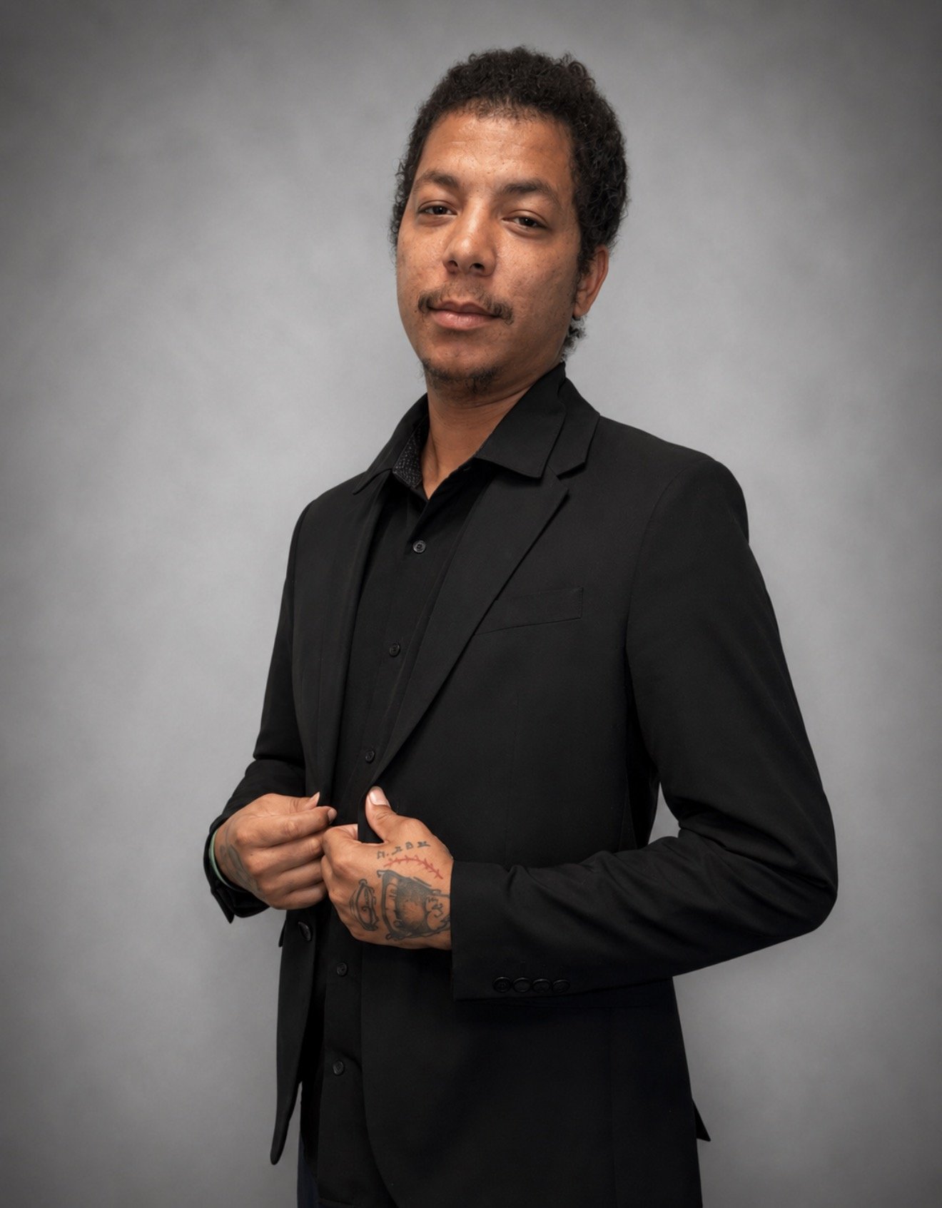 Professional picture, gray background, Multi-racial man, with tattoo hands, nice grin, in a black shirt, and black suit