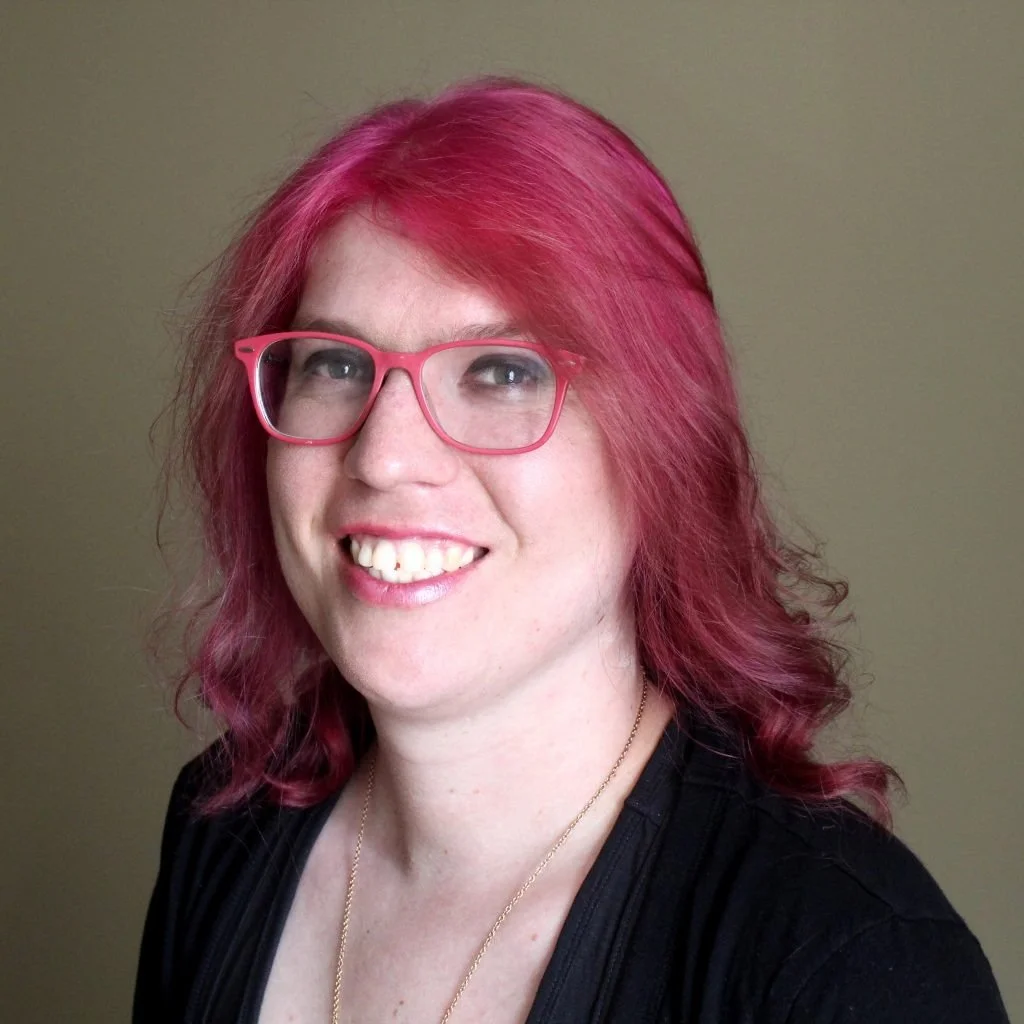 A woman with shoulder-length wavy pink hair, pink glasses, smiling, wearing a black top and a gold necklace, standing against a plain beige background.