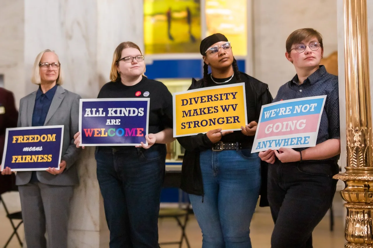 Four diverse individuals standing in a public space holding signs promoting equality and diversity. The signs read 'FREEDOM means FAIRNESS,' 'ALL KINDS ARE WELCOME,' 'DIVERSITY MAKES WV STRONGER,' and 'WE'RE NOT GOING ANYWHERE.'