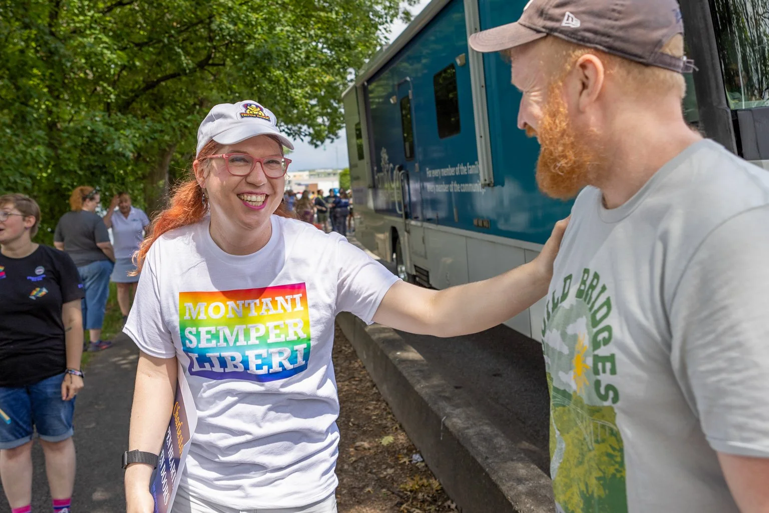 A woman with red hair, glasses, and a white cap smiling and talking to a man with a red beard and a gray cap at an outdoor event with a crowd, trees, and a blue bus in the background.