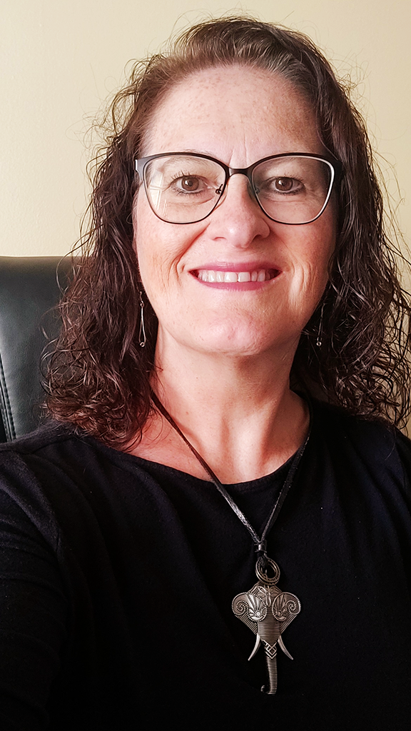 A woman with curly brown hair, glasses, and earrings, smiling in front of a beige wall, wearing a black top and a distinctive necklace with a large, ornate pendant.