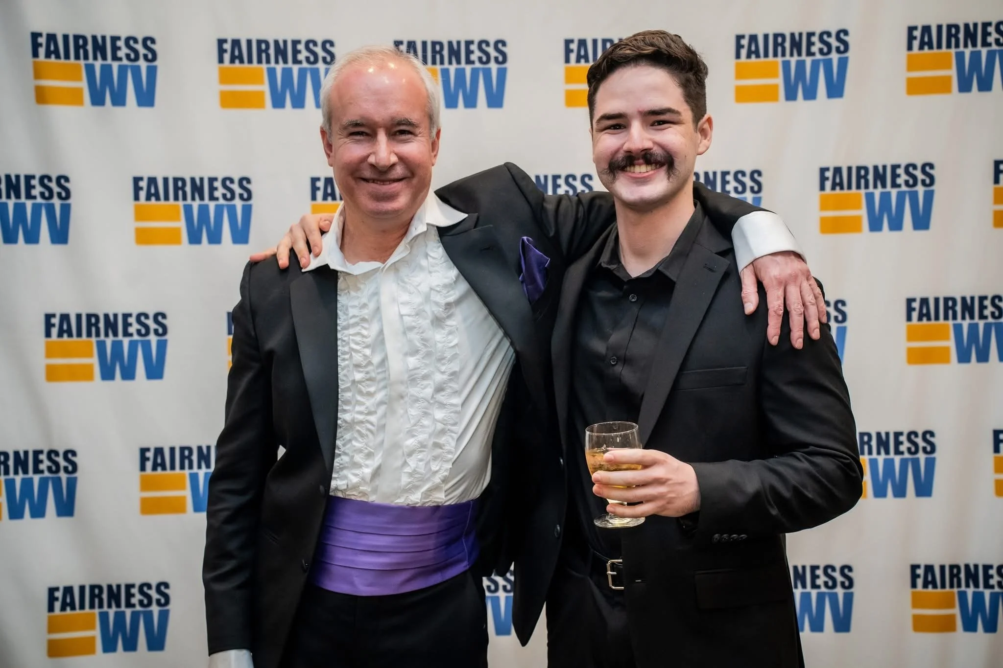 Two men dressed in tuxedos smiling with arms around each other at a Fairness West Virginia event, one holding a glass of wine.