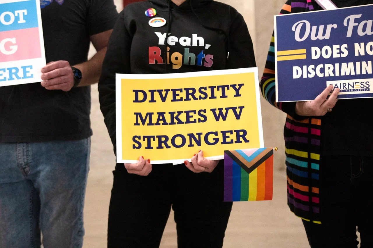 People holding signs supporting diversity and equality, including one sign that reads "Diversity Makes WV Stronger" and others advocating against discrimination and for LGBTQ+ rights.