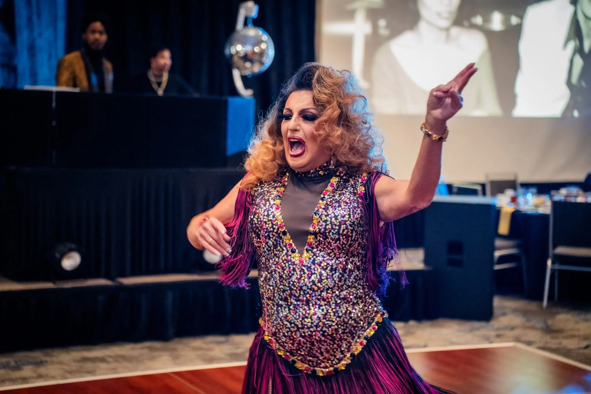 A woman with curly, blonde and brown hair wearing a shiny, colorful, sequin top and a purple fringe skirt dancing passionately indoors at a party or celebration.