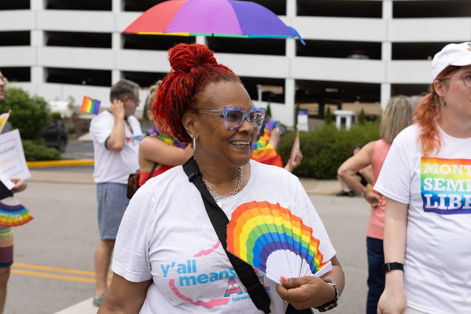 Smiling woman with red hair in dreadlocks holding a rainbow fan at a pride parade.