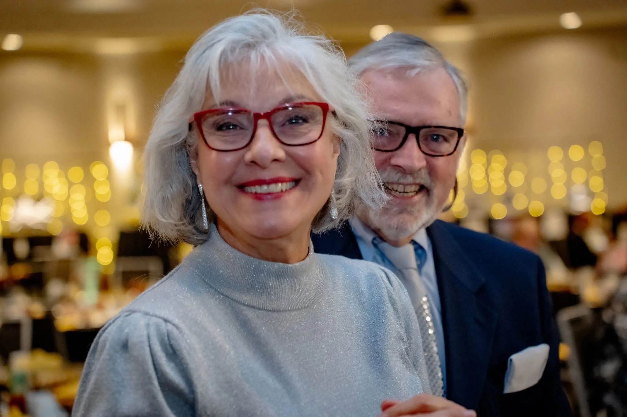 Smiling elderly woman with gray hair, red glasses, and a sparkly gray top, standing next to a smiling elderly man with gray hair, glasses, and a dark suit, in a decorated indoor setting with warm lighting and blurred background.