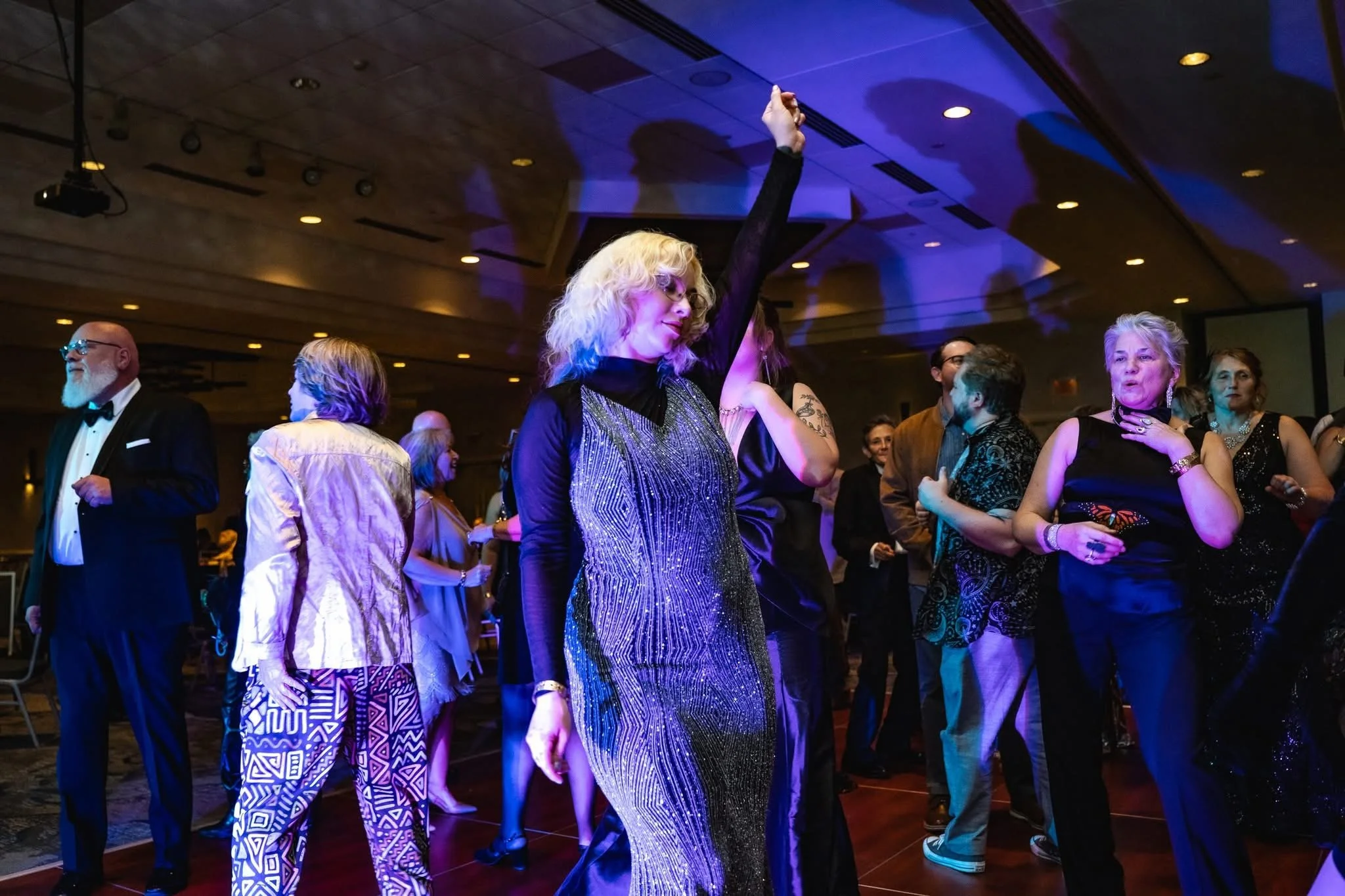 People dancing and socializing at an indoor event with dim lighting and colorful spotlights.
