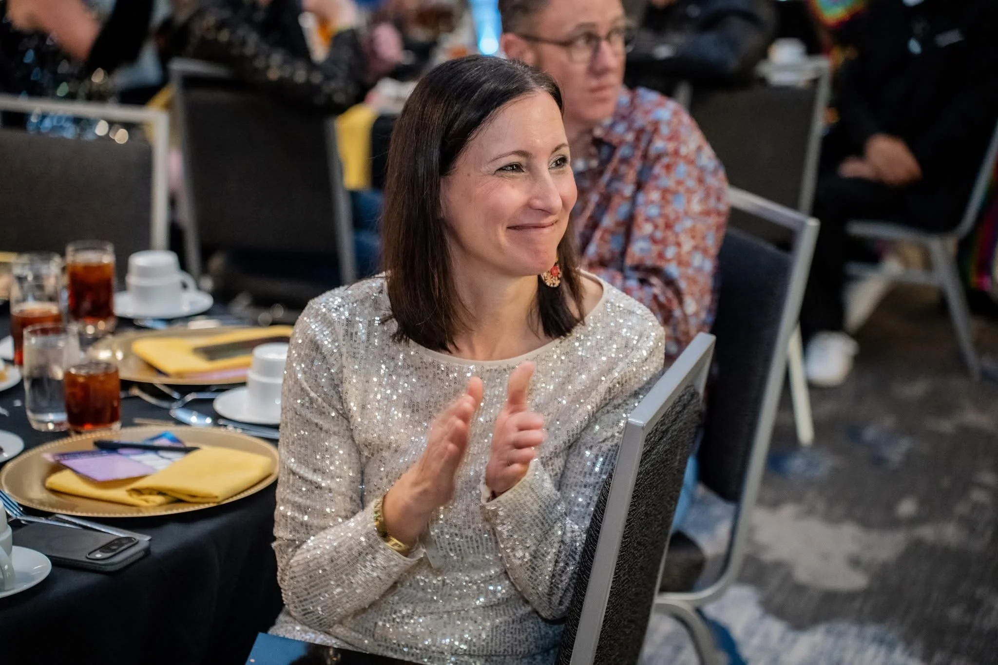 A woman in a shiny, sequined top sitting at a table, clapping and smiling at an event.