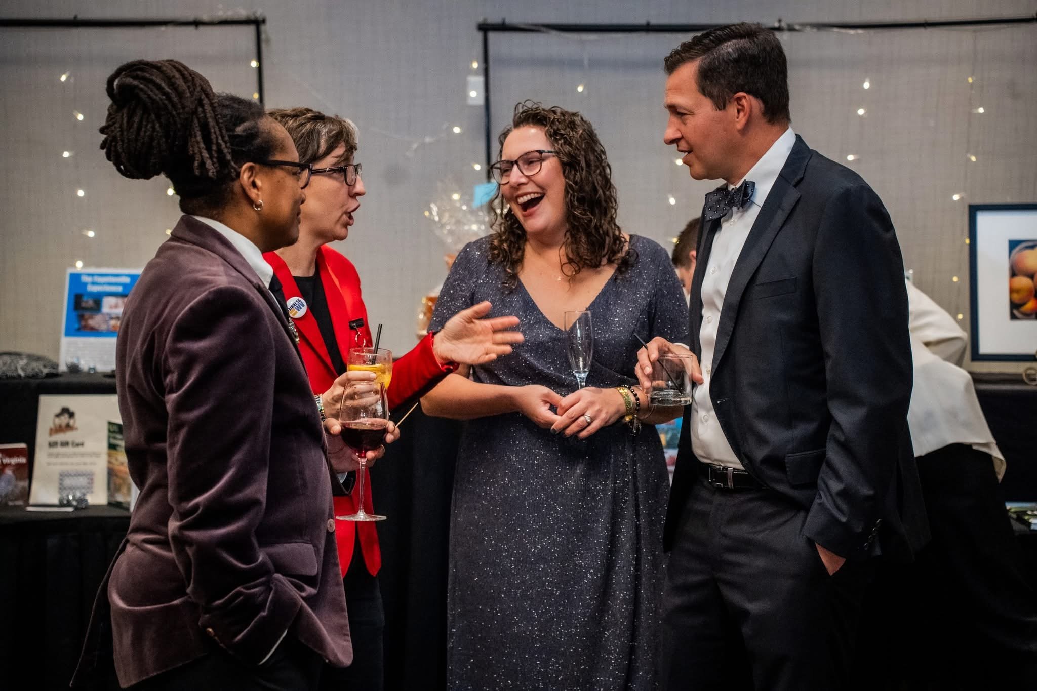 Four people in formal attire having a conversation at a social event, holding drinks, with a decorated background.