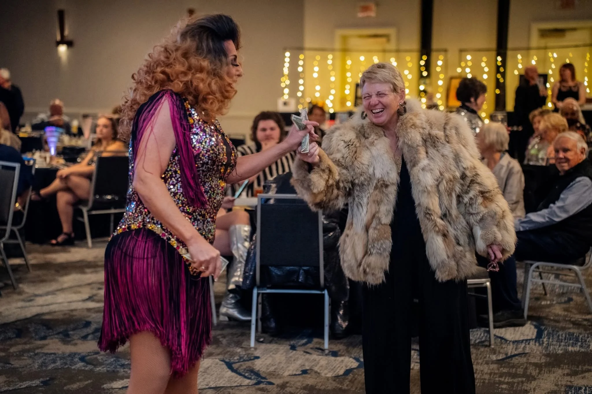 Two women smiling and exchanging money at a social event, one wearing a colorful, sequined dress with fringe and the other wearing a fur coat, in a decorated banquet hall with seated guests in the background.