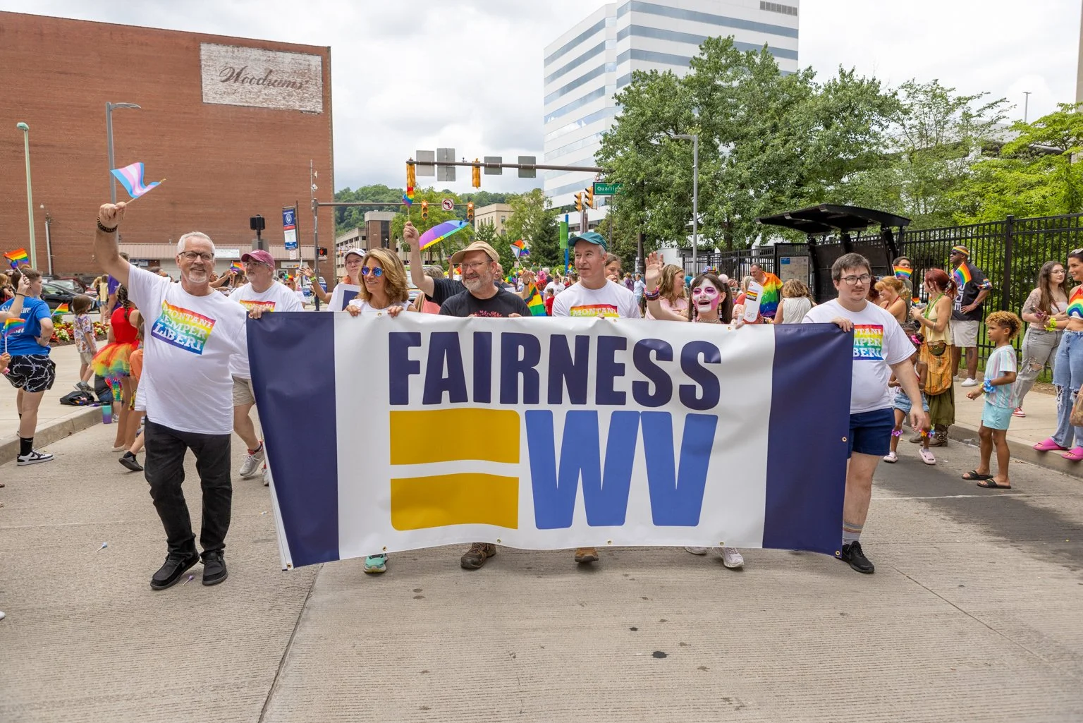 Group of people holding a banner that reads 'FAIRNESS WV' at a pride parade, with many participants and spectators in the background, some wearing rainbow accessories and face paint.