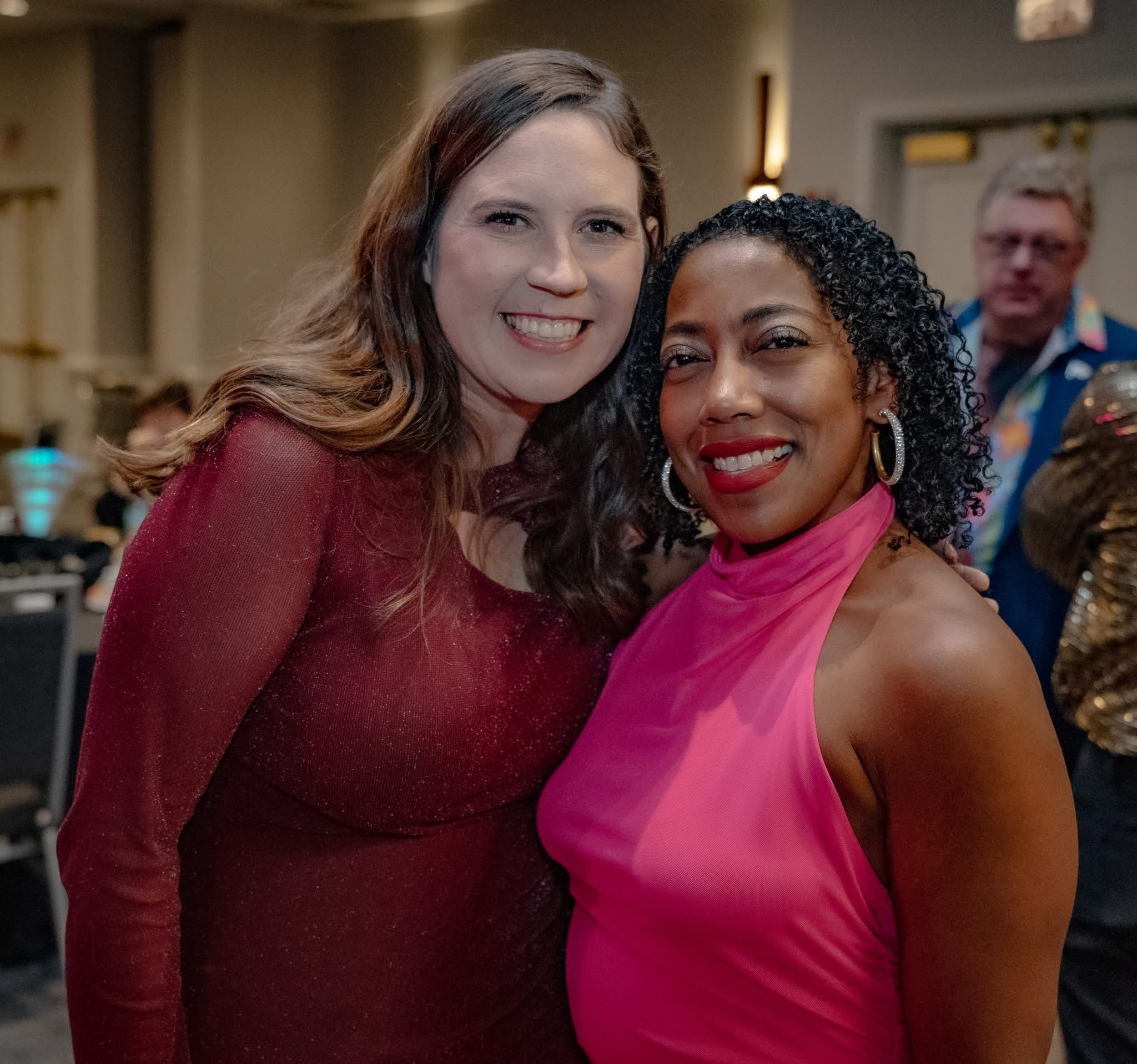Two women smiling at an indoor social event, one wearing a burgundy dress, the other in a pink halter top, with a man in glasses in the background.
