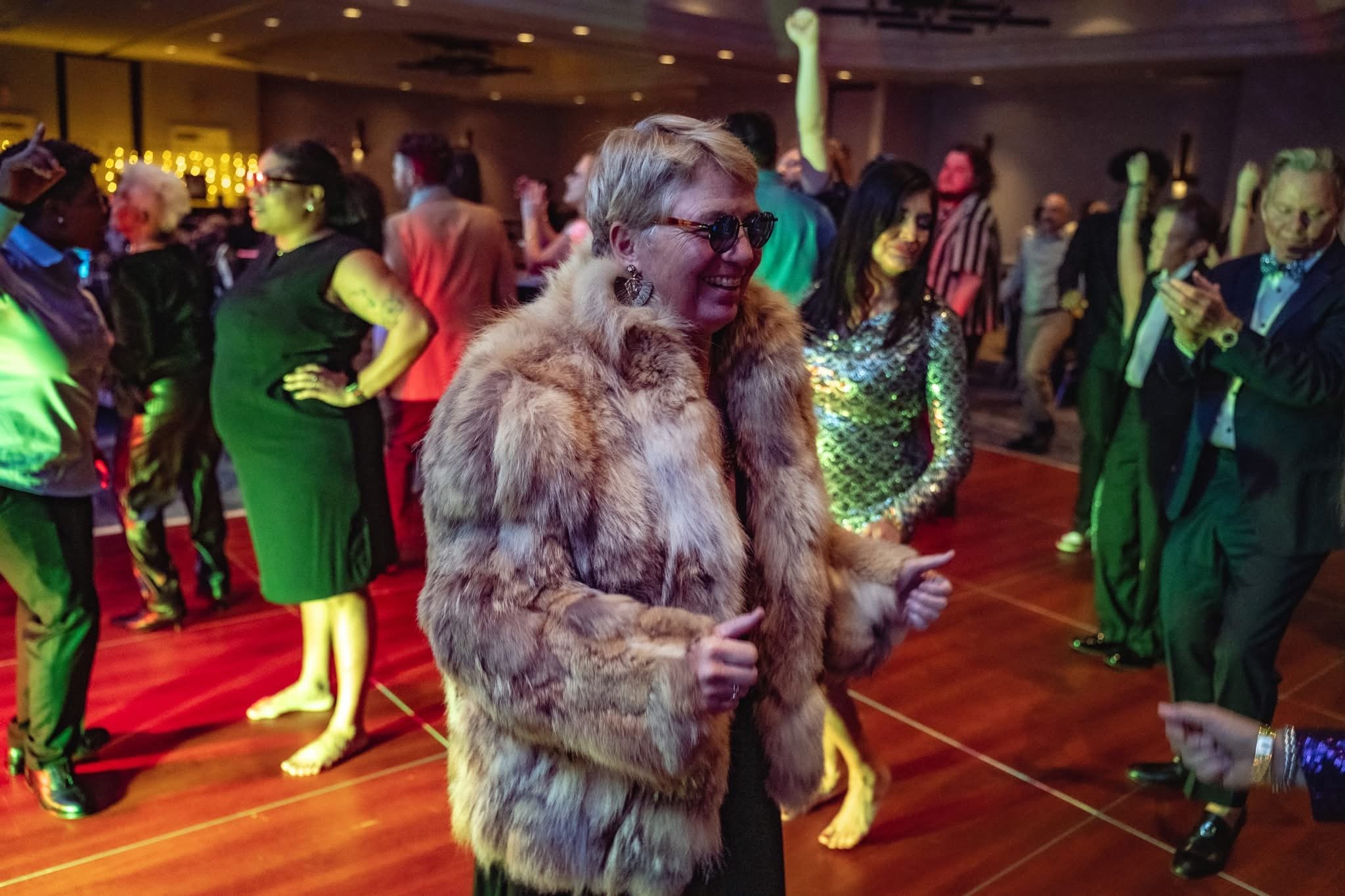 Woman wearing a fur coat and sunglasses dancing at a party with many people in the background.