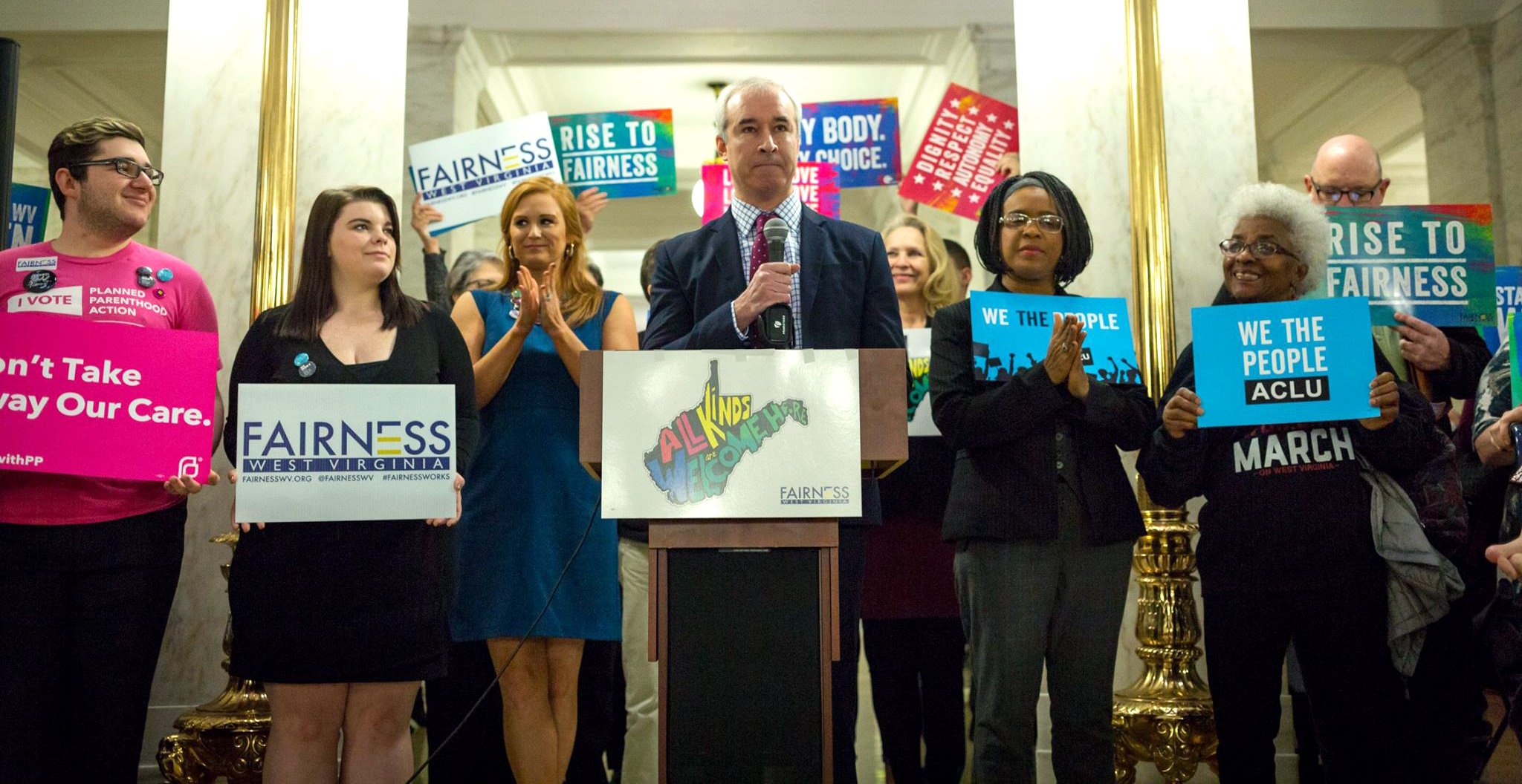 Group of diverse people at a rally or protest holding signs with messages like "Fairness West Virginia" and "Rise to Fairness," standing around a man at a podium speaking into a microphone, in a formal setting with gold accents and mirrors on the wall.