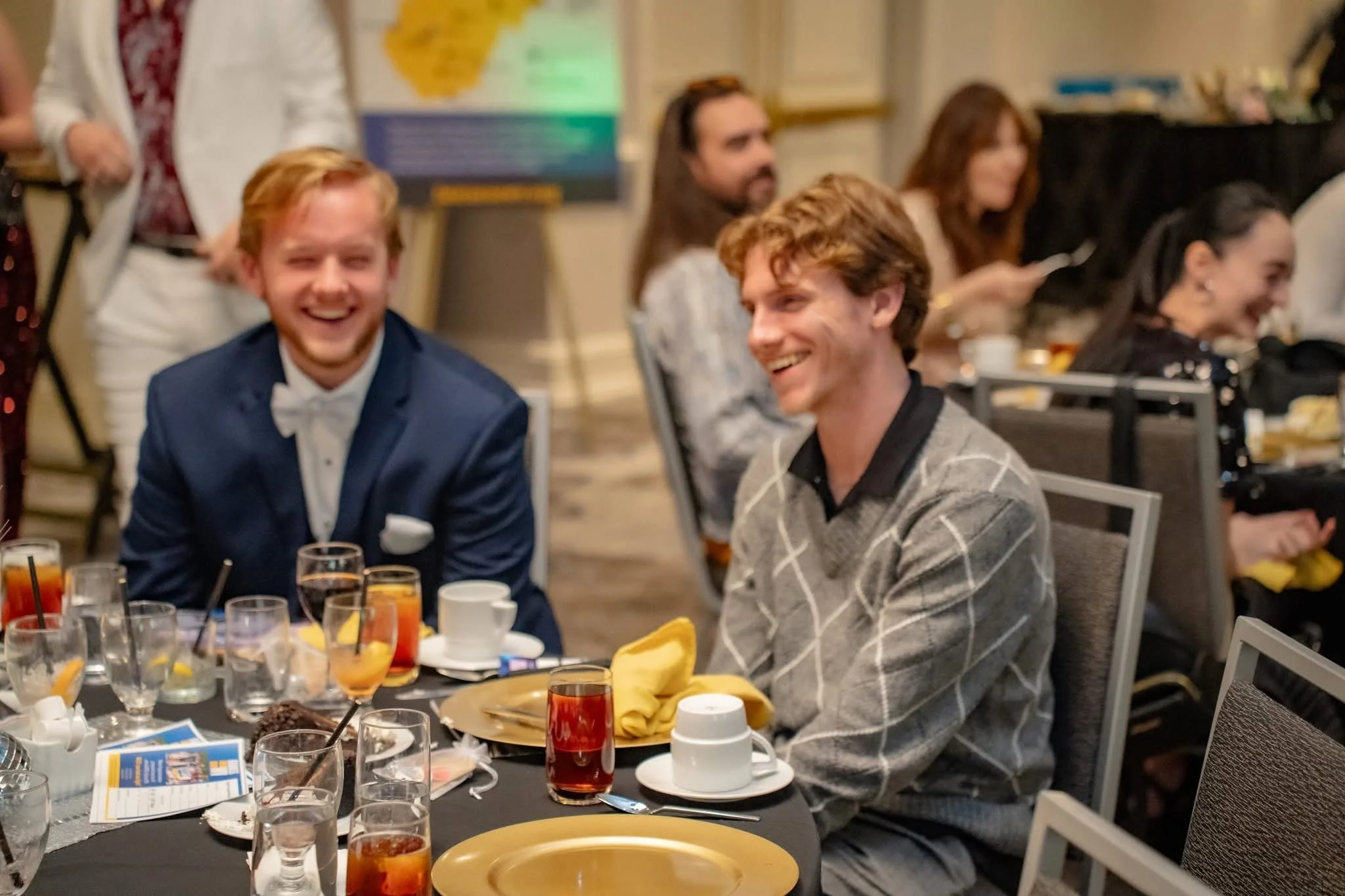 Two men are sitting at a table during a celebration or formal event, smiling and laughing. The man on the left is in a navy suit and bow tie, while the man on the right is in a gray sweater with a diamond pattern. The table has drinks, plates, and na