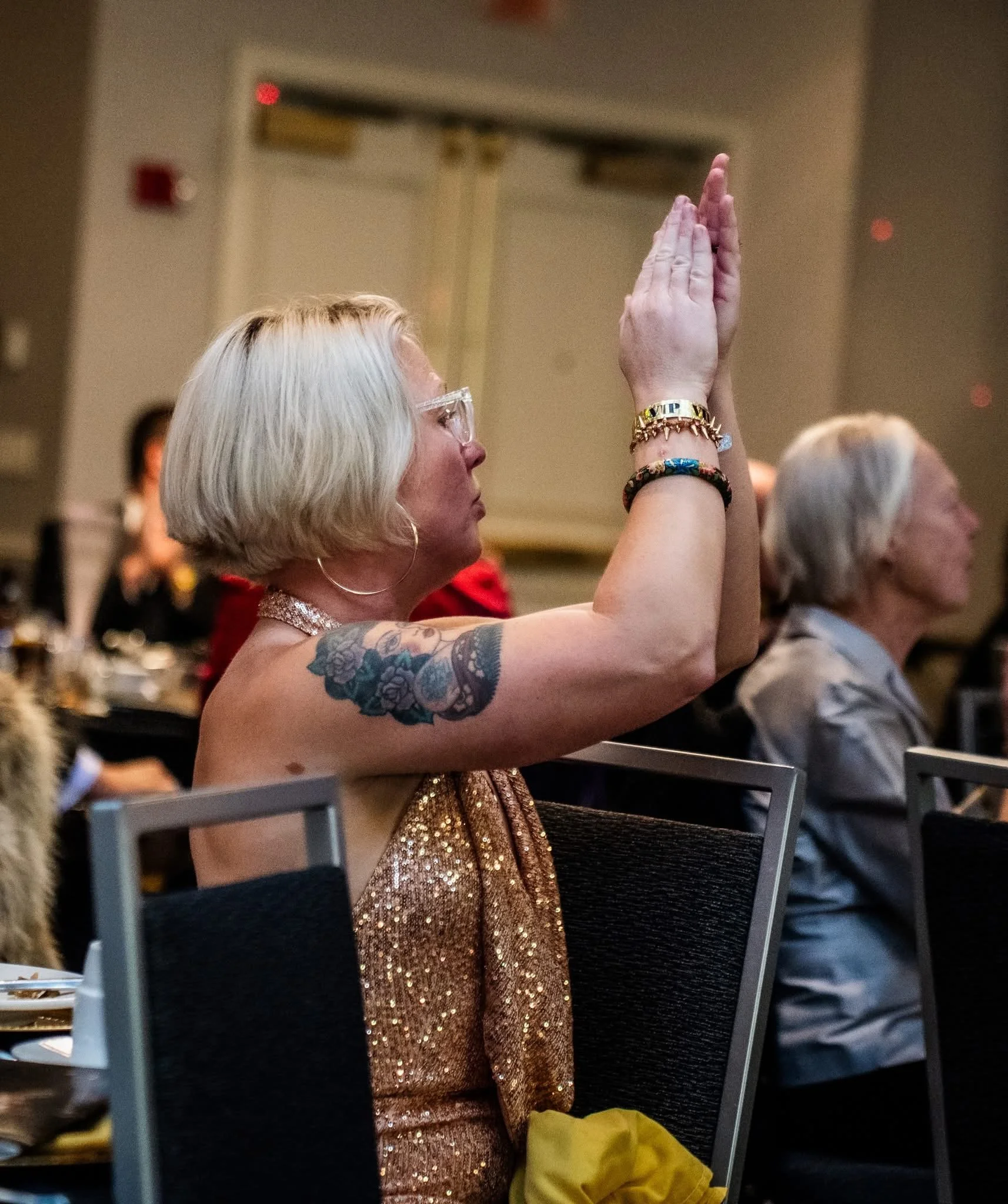 A woman with short blonde hair, glasses, and tattoos on her arm is seated, wearing a sparkly dress, with her hands clasped together in a prayer or worship position during an event.