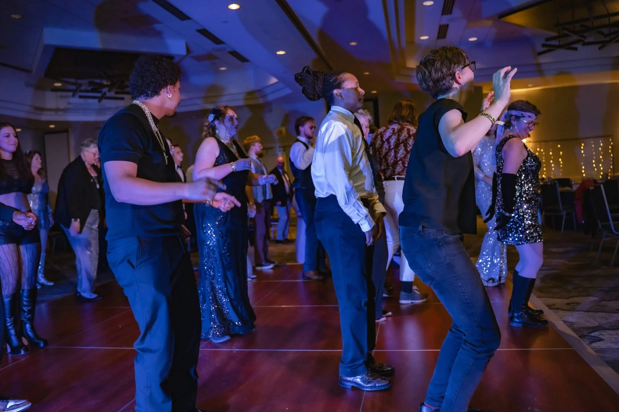 People dancing at an event indoors under colorful lighting, with some wearing shiny or fashionable attire.