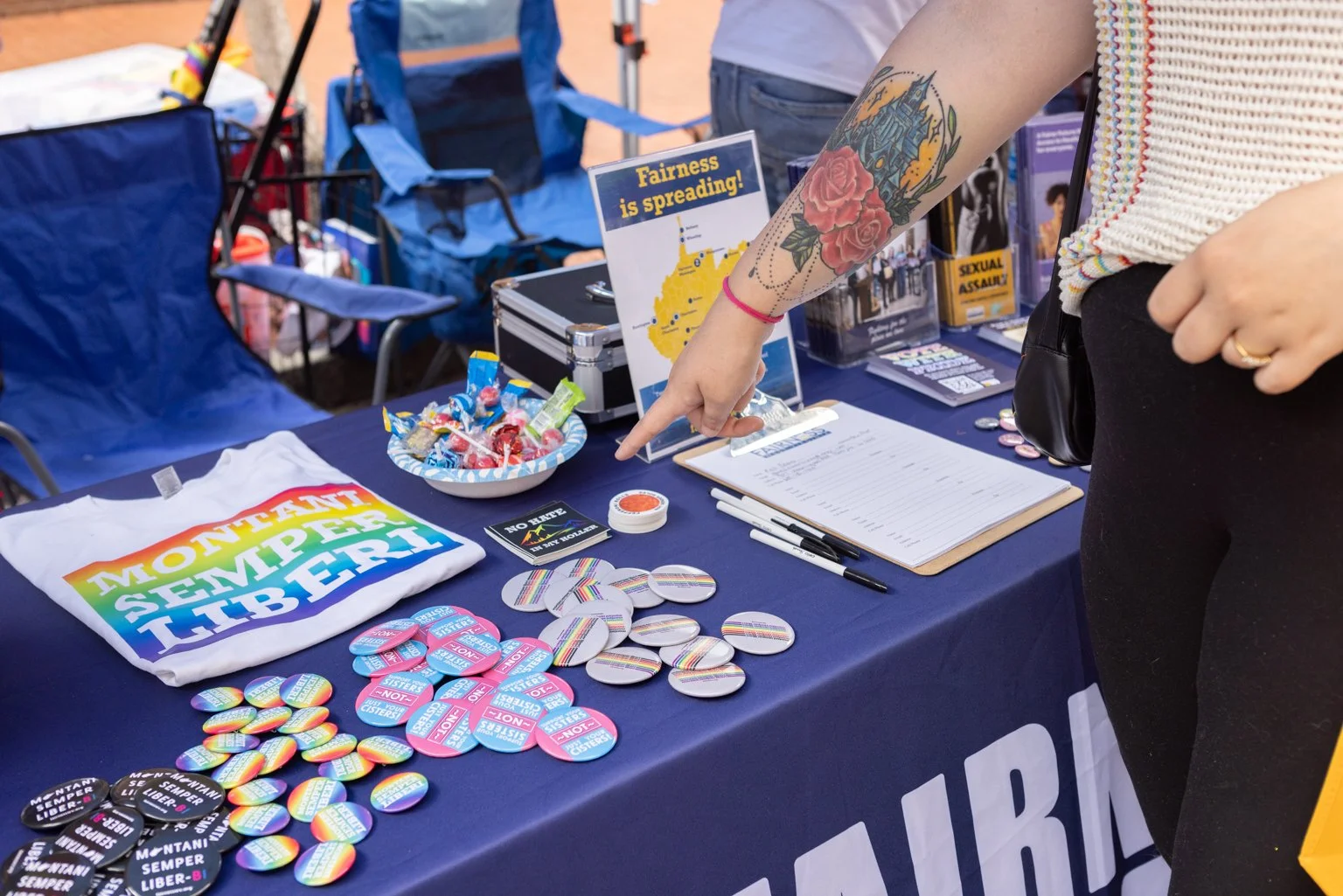 A person at a booth points at a sign that reads "Fairness is spreading!" with a map of Montana. The table displays buttons and pins with messages supporting reproductive rights, including rainbow-themed buttons, and informational materials about reproductive justice.