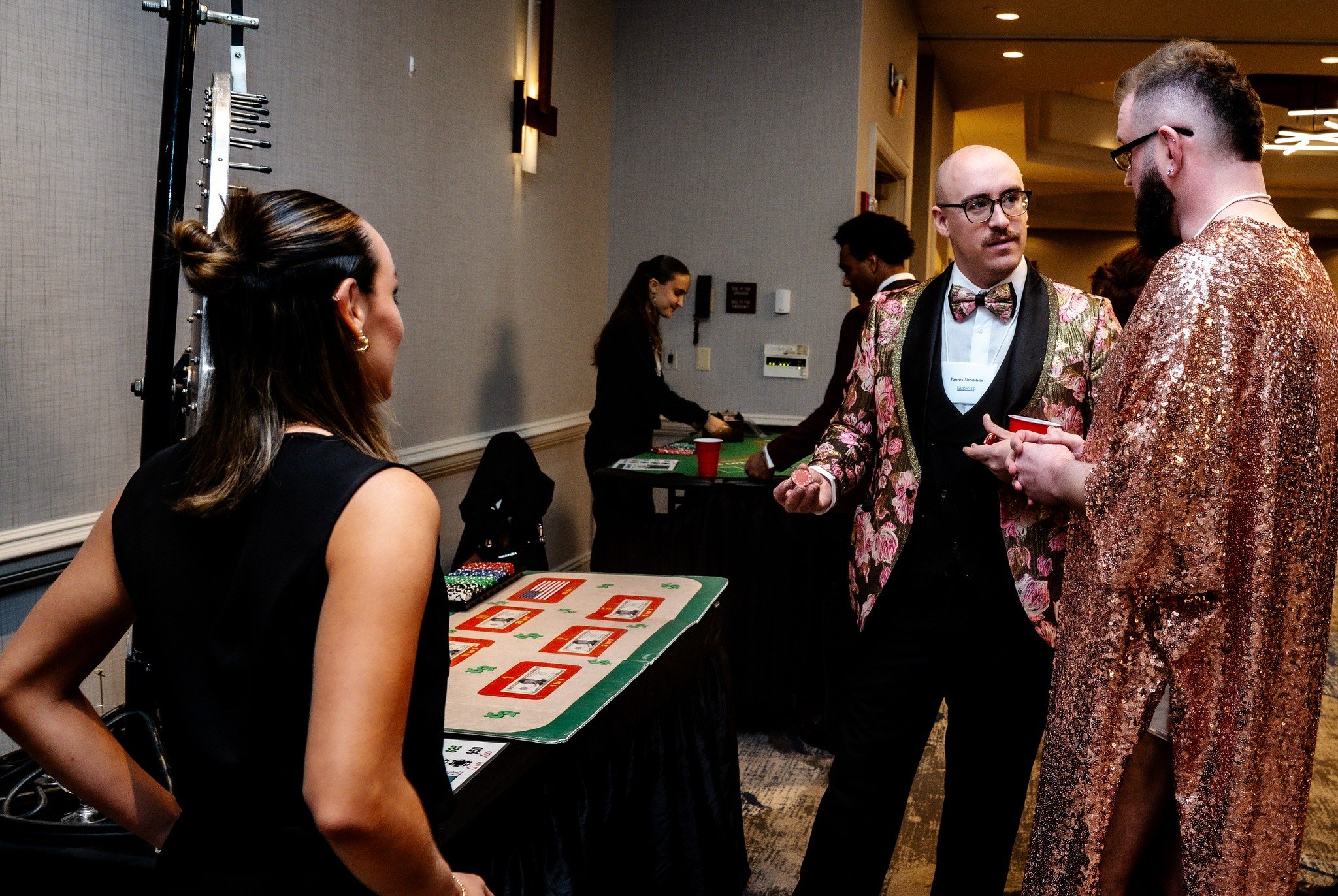 People socializing at an event, some dressed in colorful, shiny coats, with tables and games in the background.