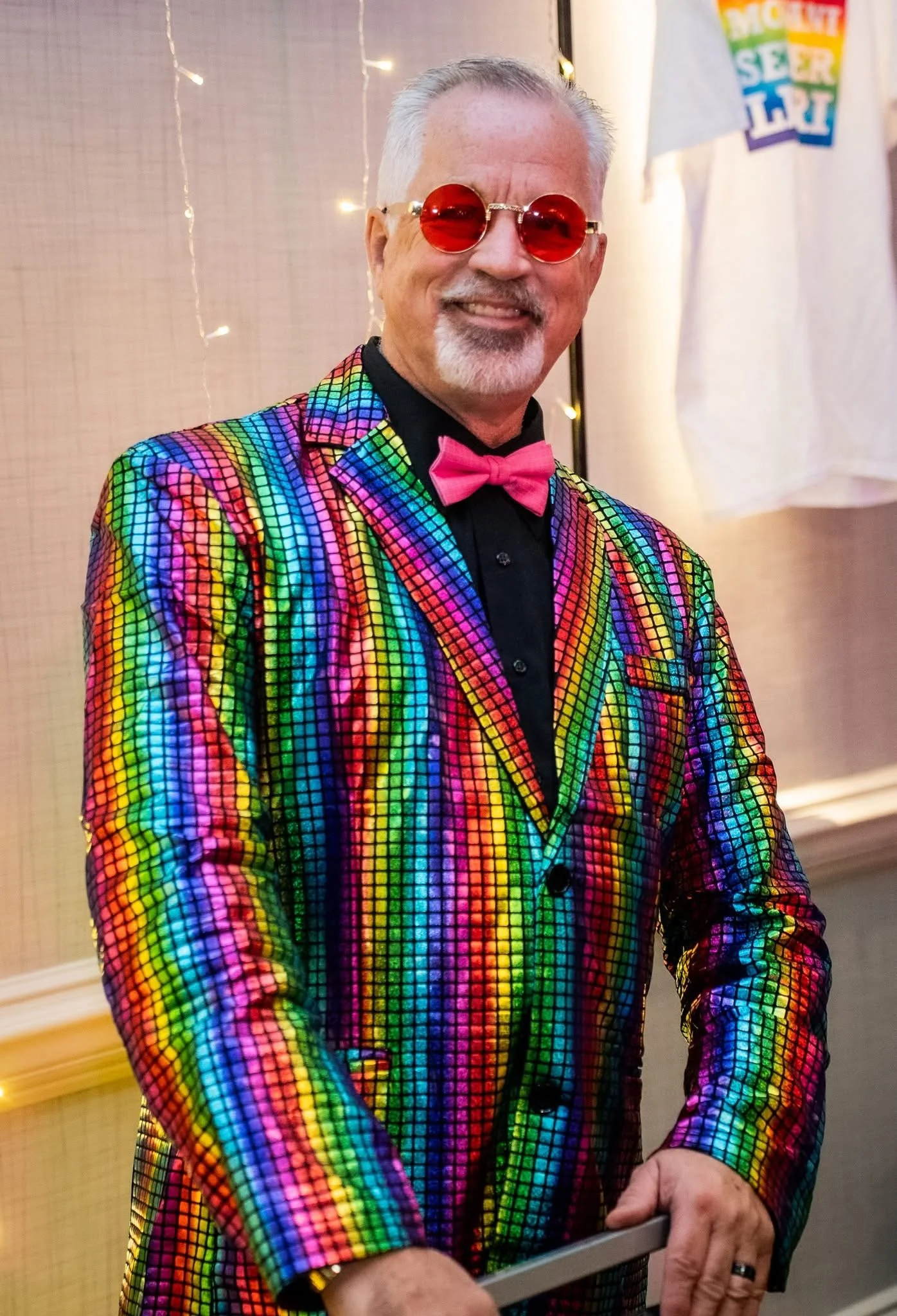 A smiling man dressed in a colorful, pixelated rainbow blazer with a pink bow tie, round red sunglasses, and a black shirt.