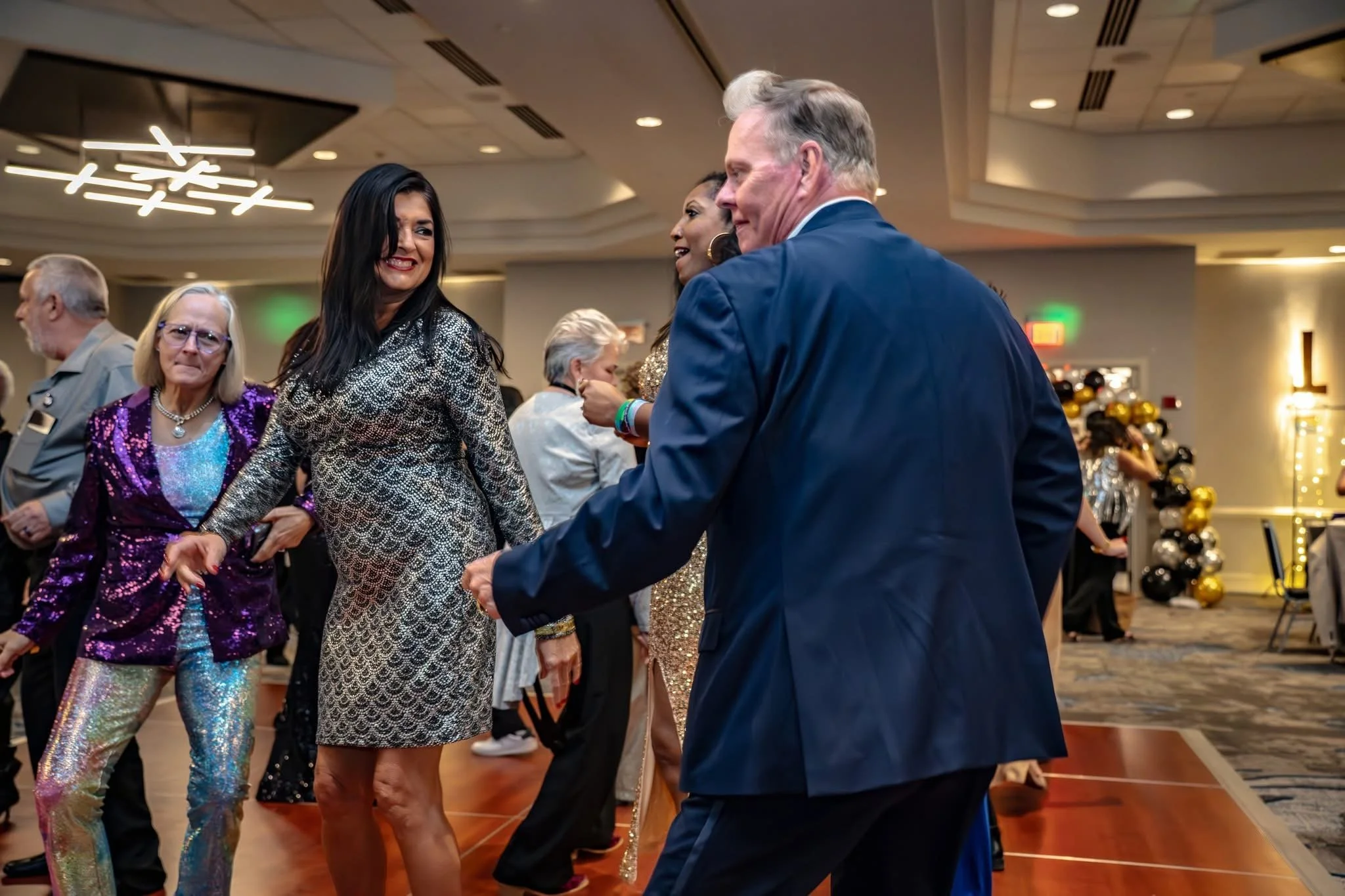 People dancing and smiling at a party or celebration indoors, with balloons and decorations in the background.