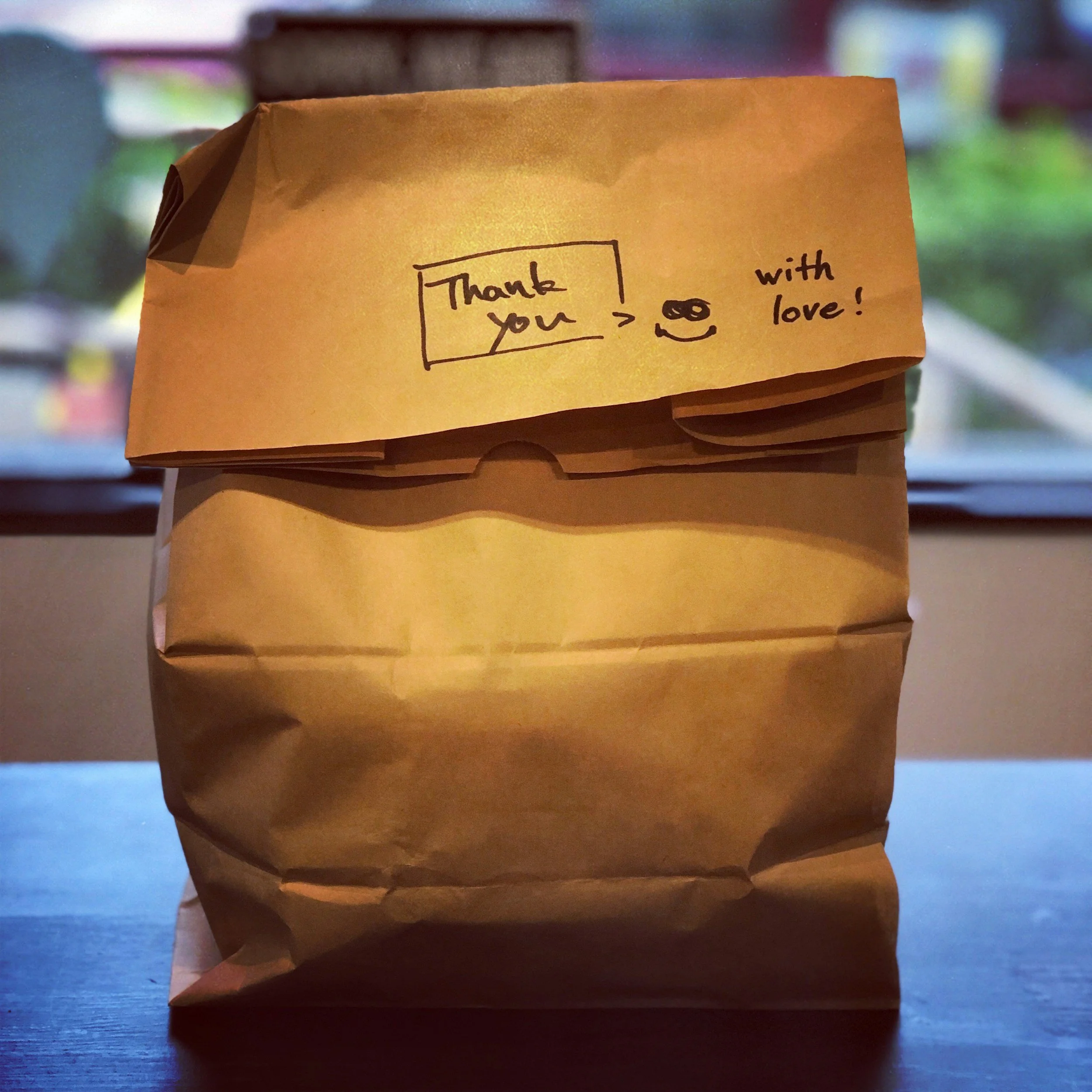 A brown paper bag with a handwritten note on it that says, 'Thank you > with love!', accompanied by a small smiley face and a pair of glasses illustration. The bag is placed on a wooden surface with a blurred background.
