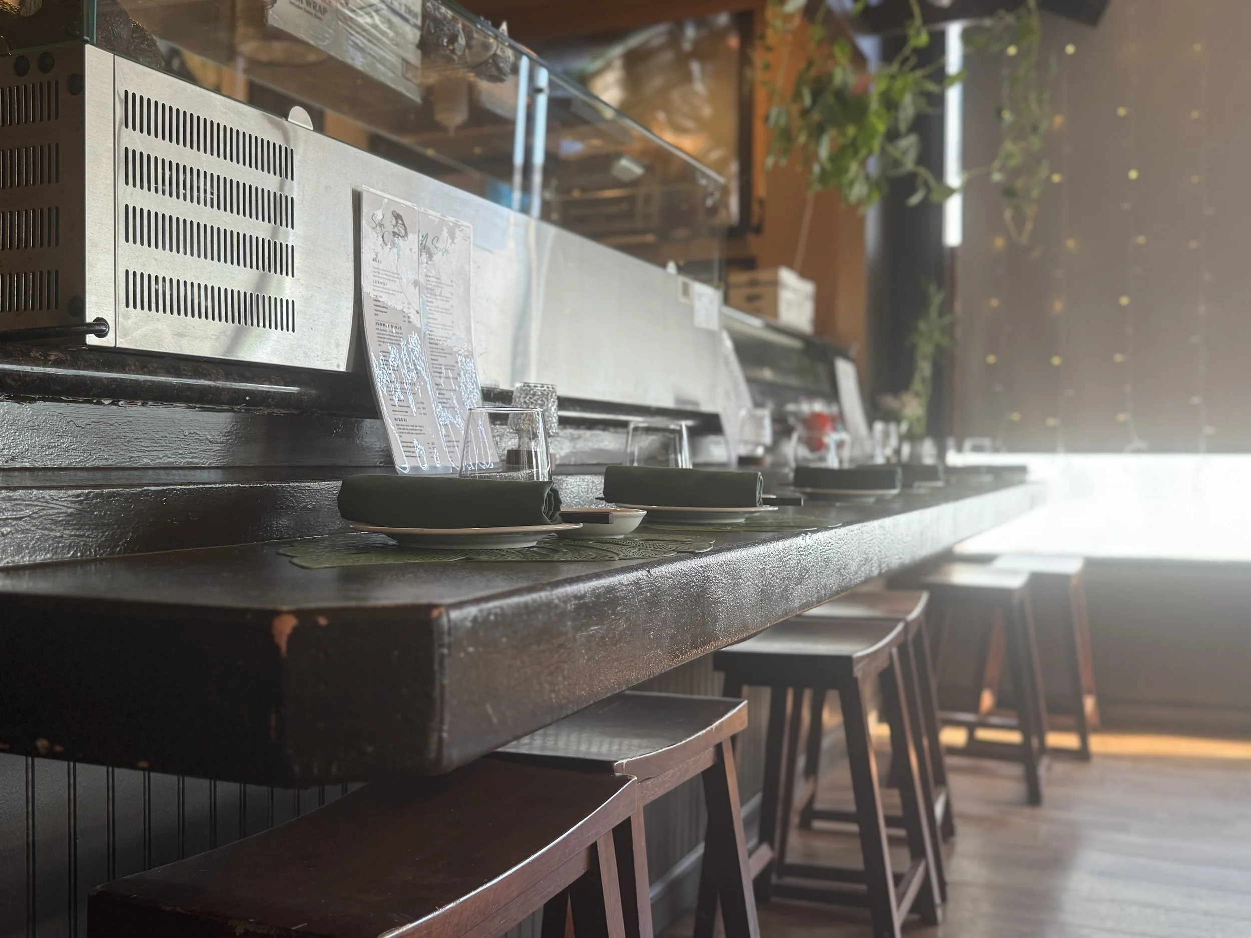 Empty restaurant counter with bar stools, menus, and place settings, with sunlight coming through large windows and plants in the background.