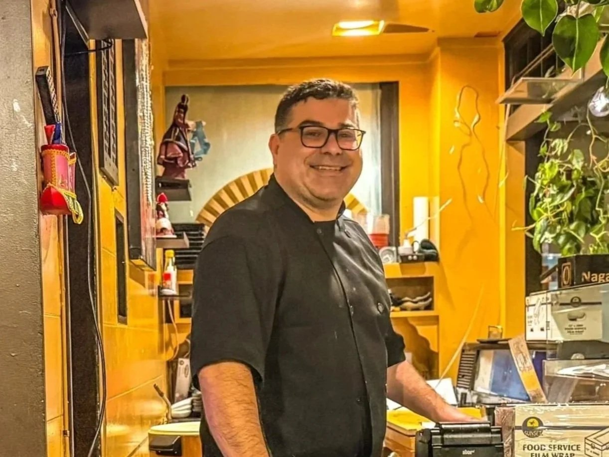 A man in a black chef's jacket smiling in a warmly lit, cozy restaurant kitchen with yellow walls, shelves with plants, and various kitchen items.