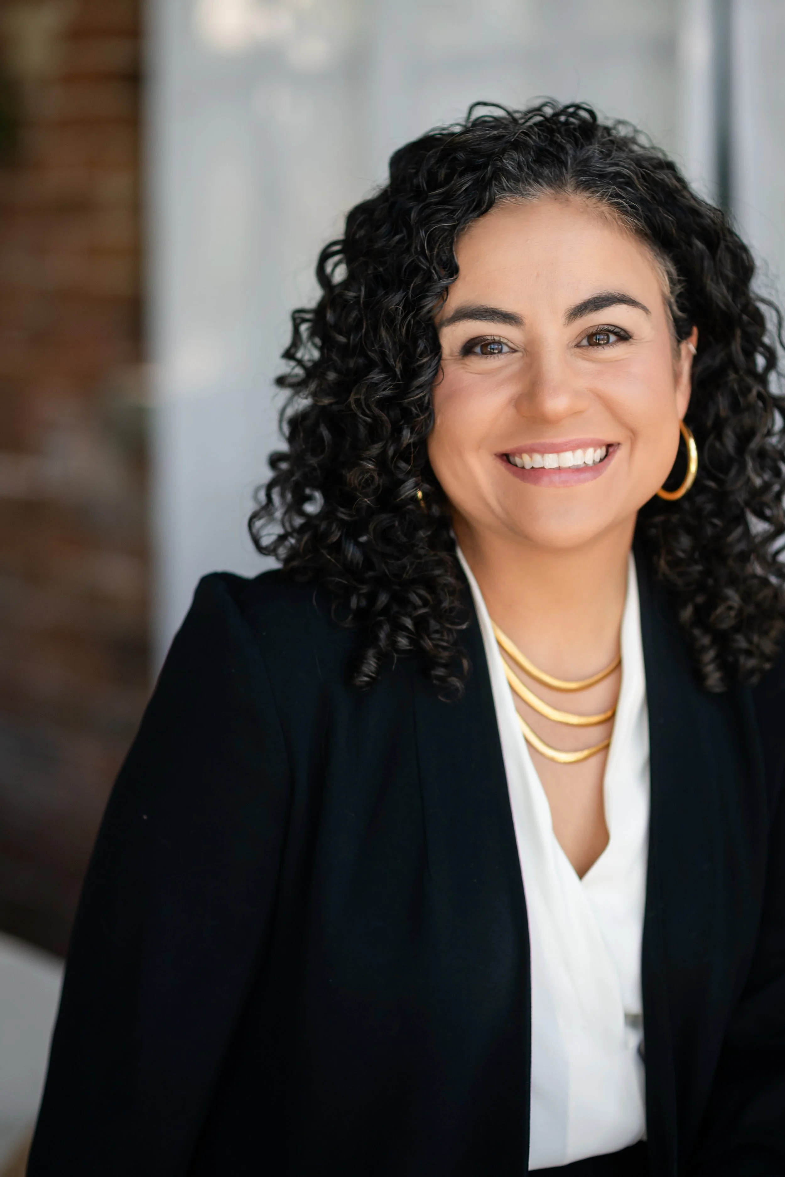 A woman with curly black hair, wearing gold hoop earrings, a gold necklace, a white blouse, and a black blazer smiles at the camera.